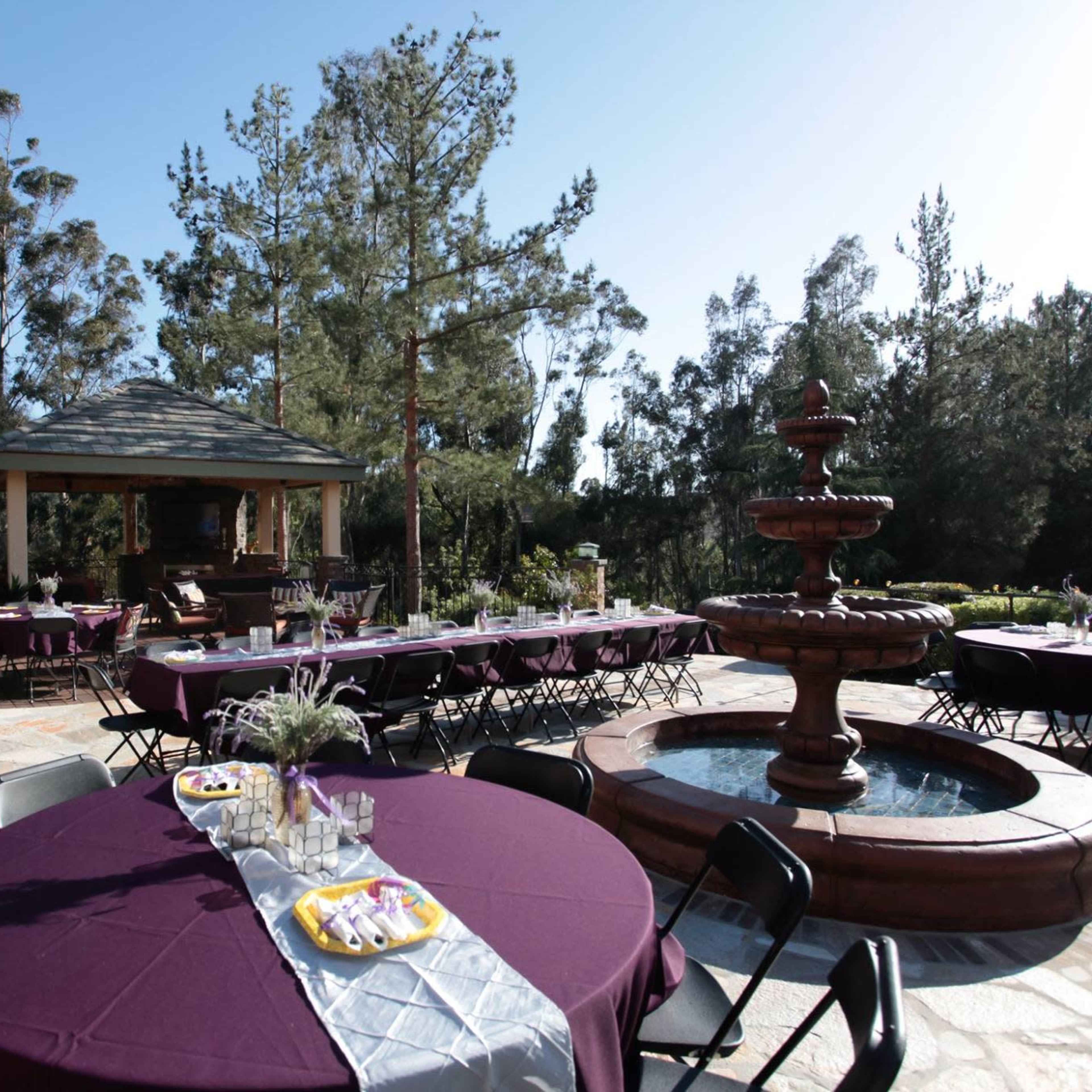 A scenic outdoor event space features purple tablecloths on round tables, a central fountain, and a gazebo surrounded by trees.