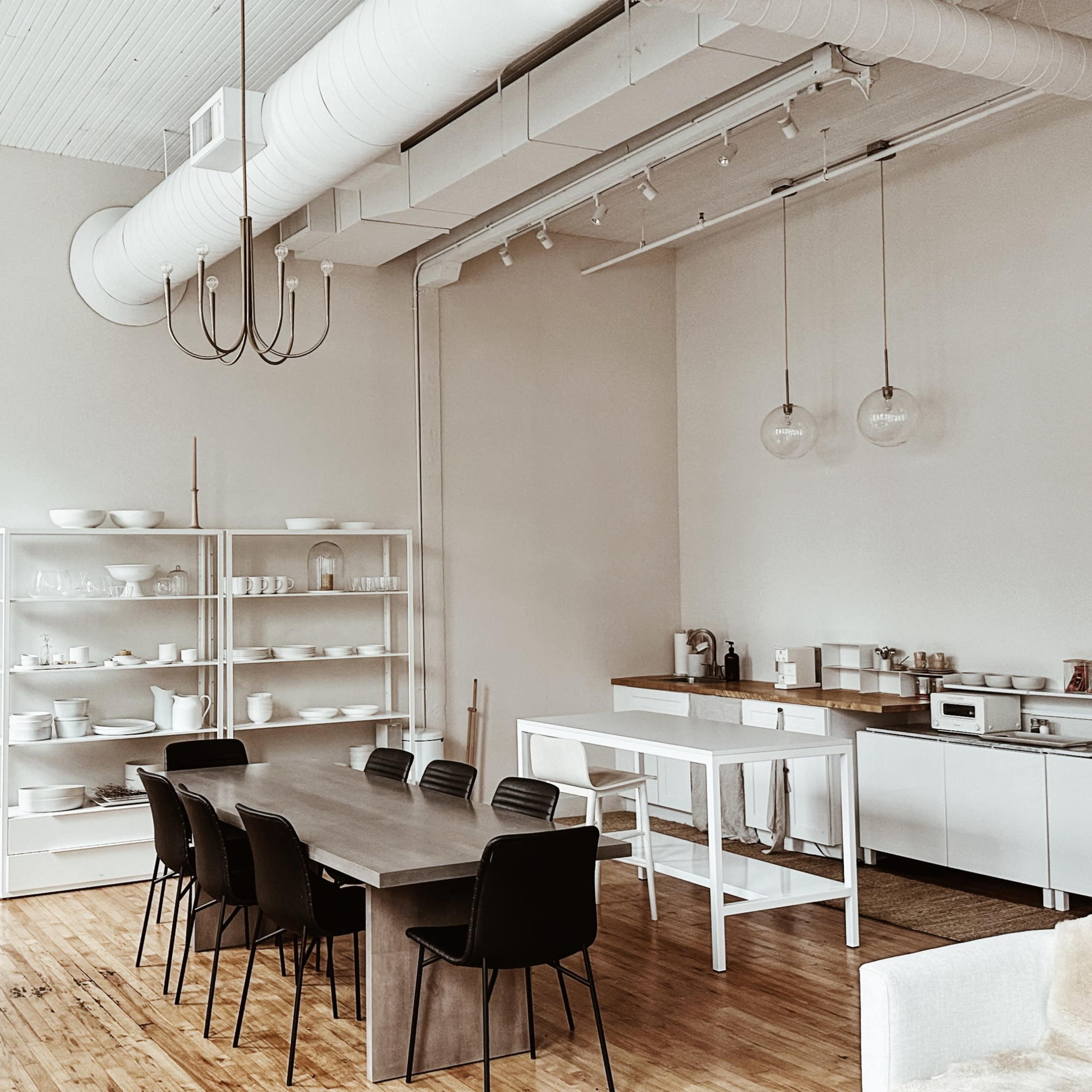 A minimalist kitchen and dining area with a long wooden table, black chairs, and shelves filled with white dishware.