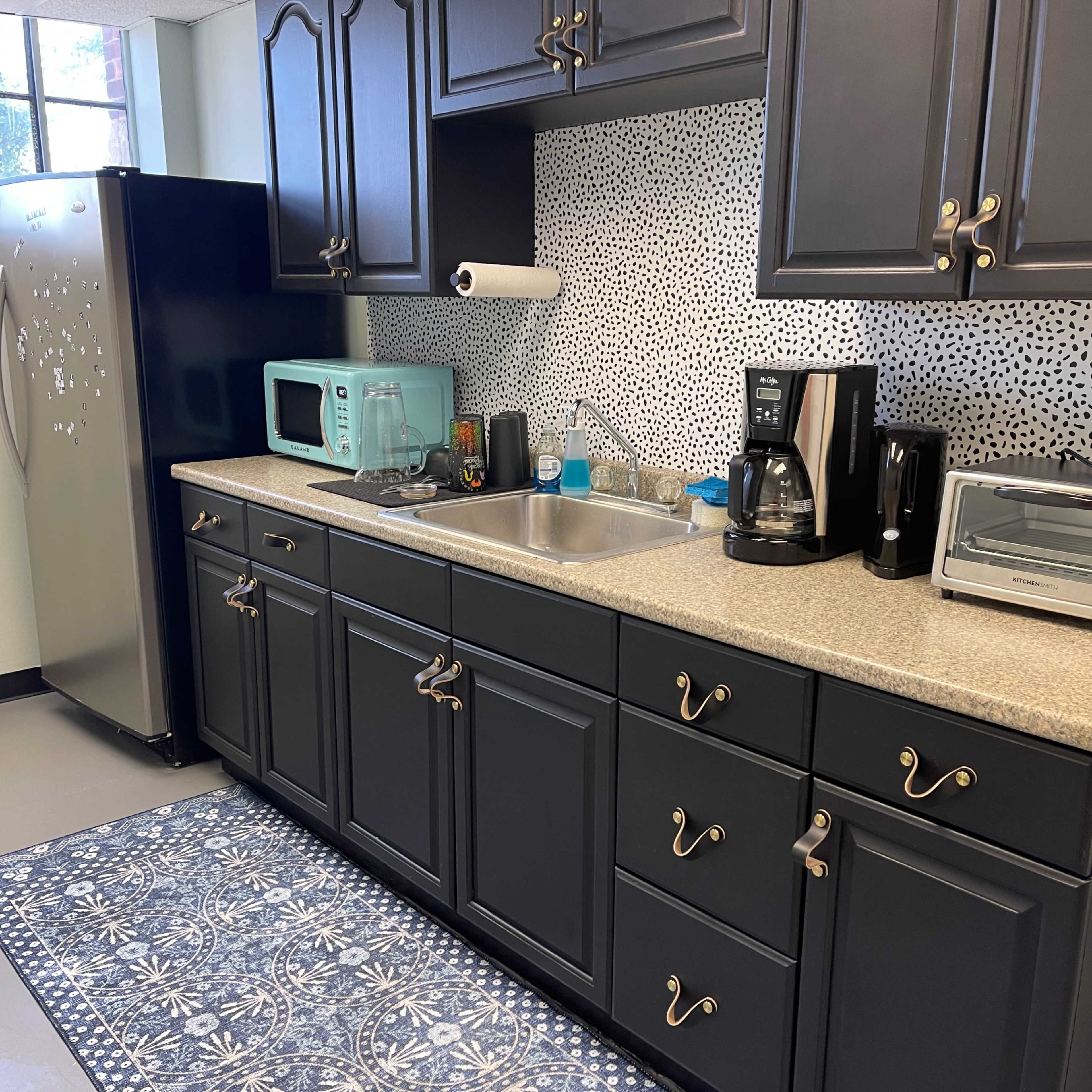 A modern kitchen with dark cabinetry, a stainless steel refrigerator, and a countertop with various appliances, including a coffee maker and microwave.