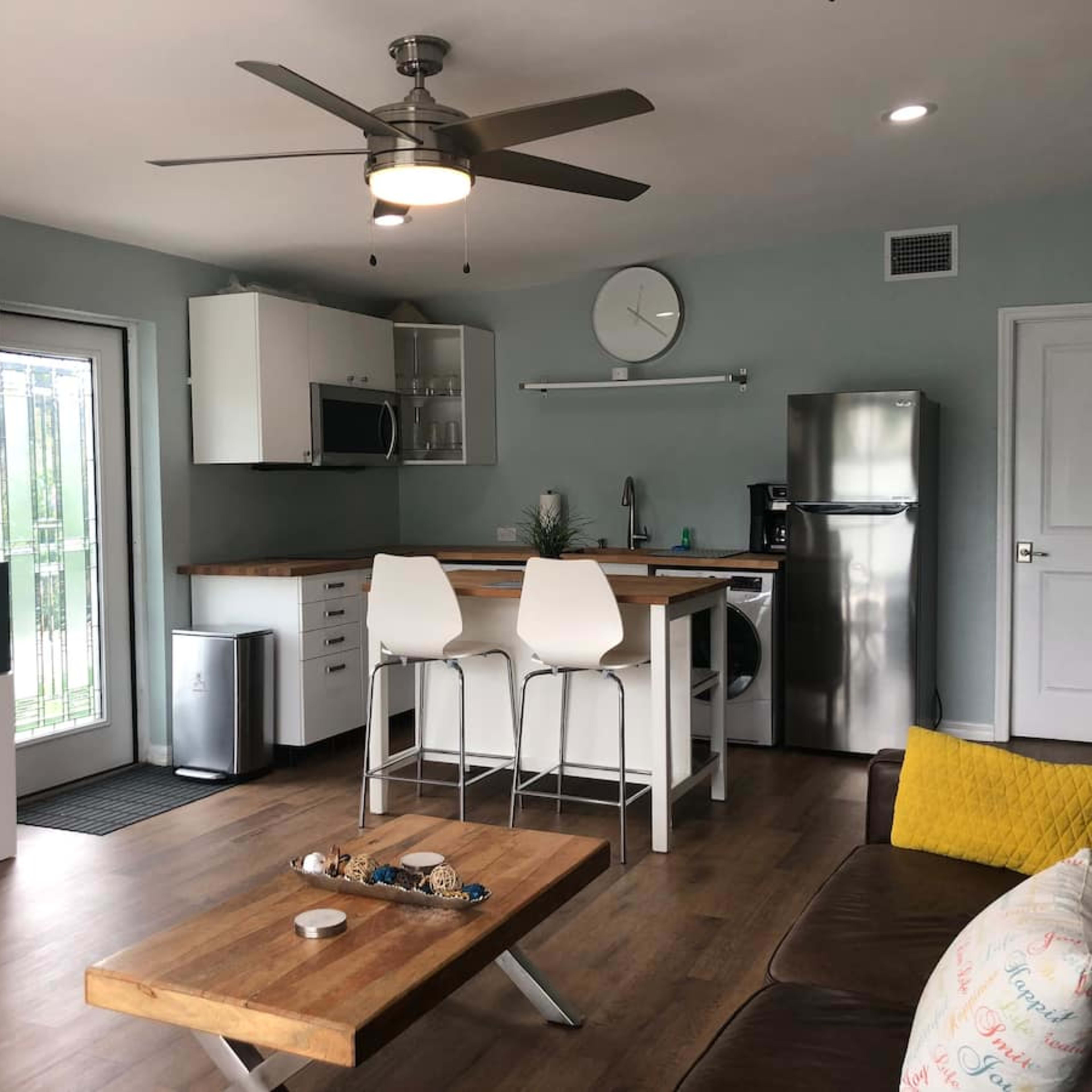 A modern kitchen and living area with a wooden table, white cabinetry, stainless steel appliances, and a ceiling fan.
