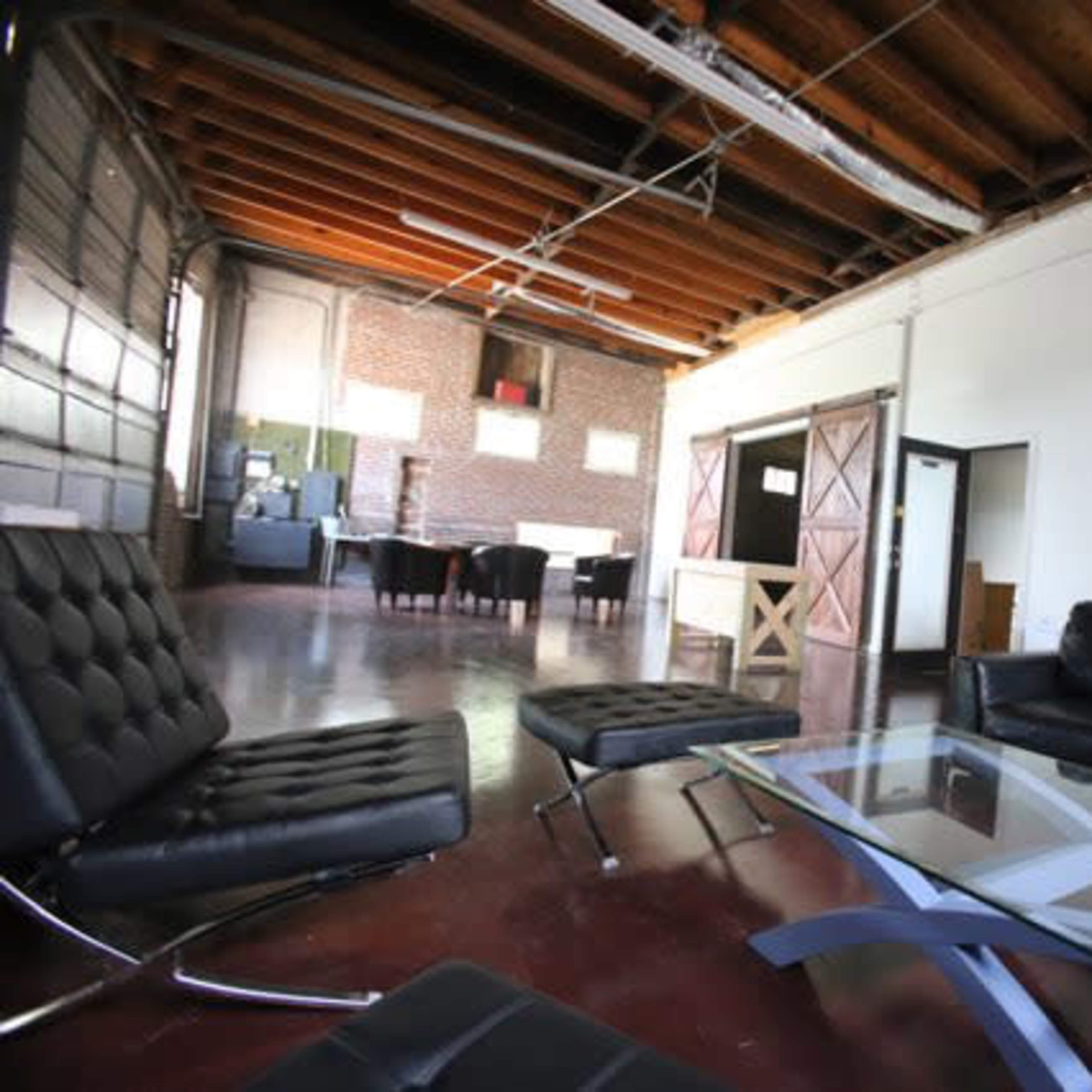 A spacious, modern living area with a black leather sofa, glass coffee table, and open layout, featuring exposed wooden beams and large windows.