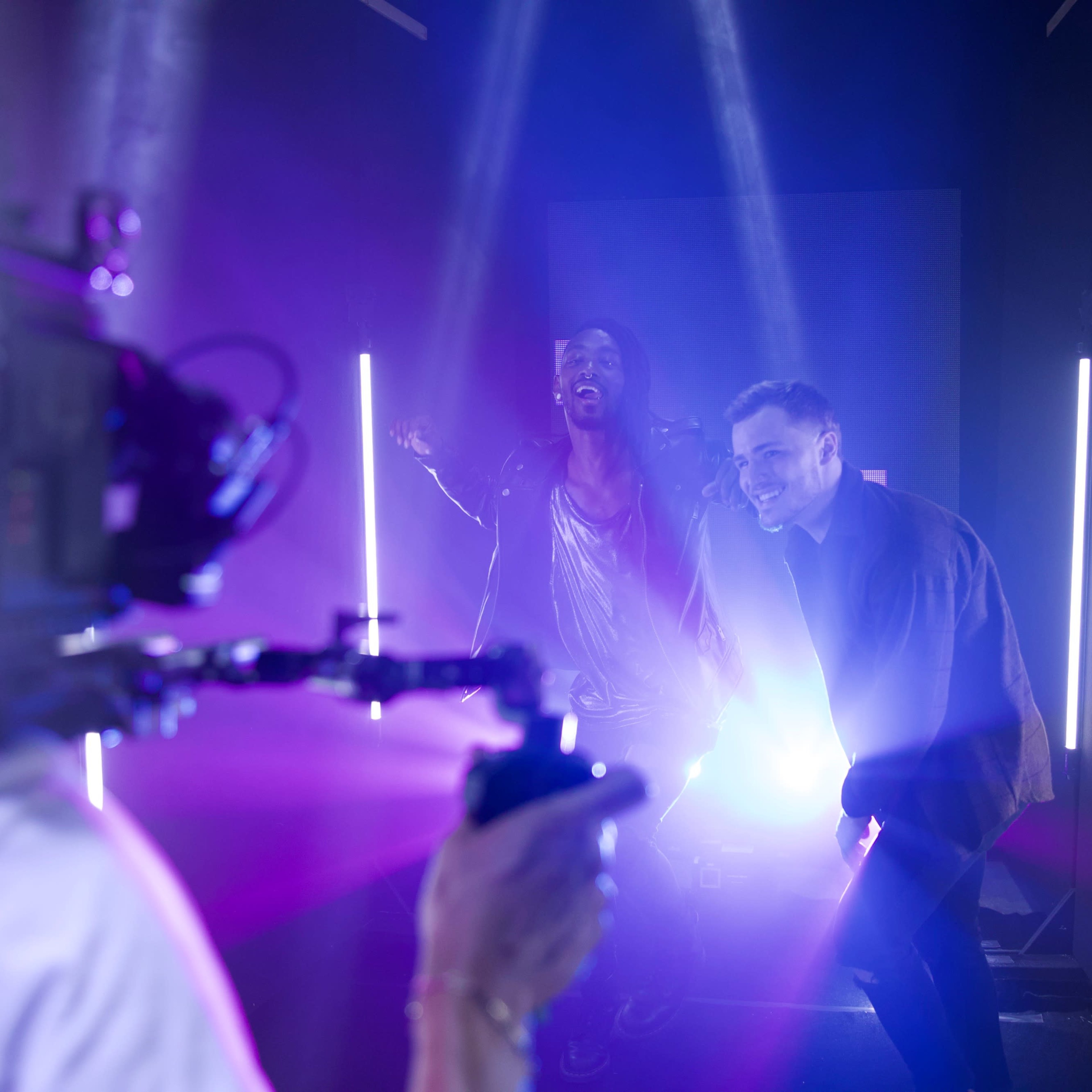 Two men perform on a stage illuminated by colorful lights while a camera operator captures the scene.