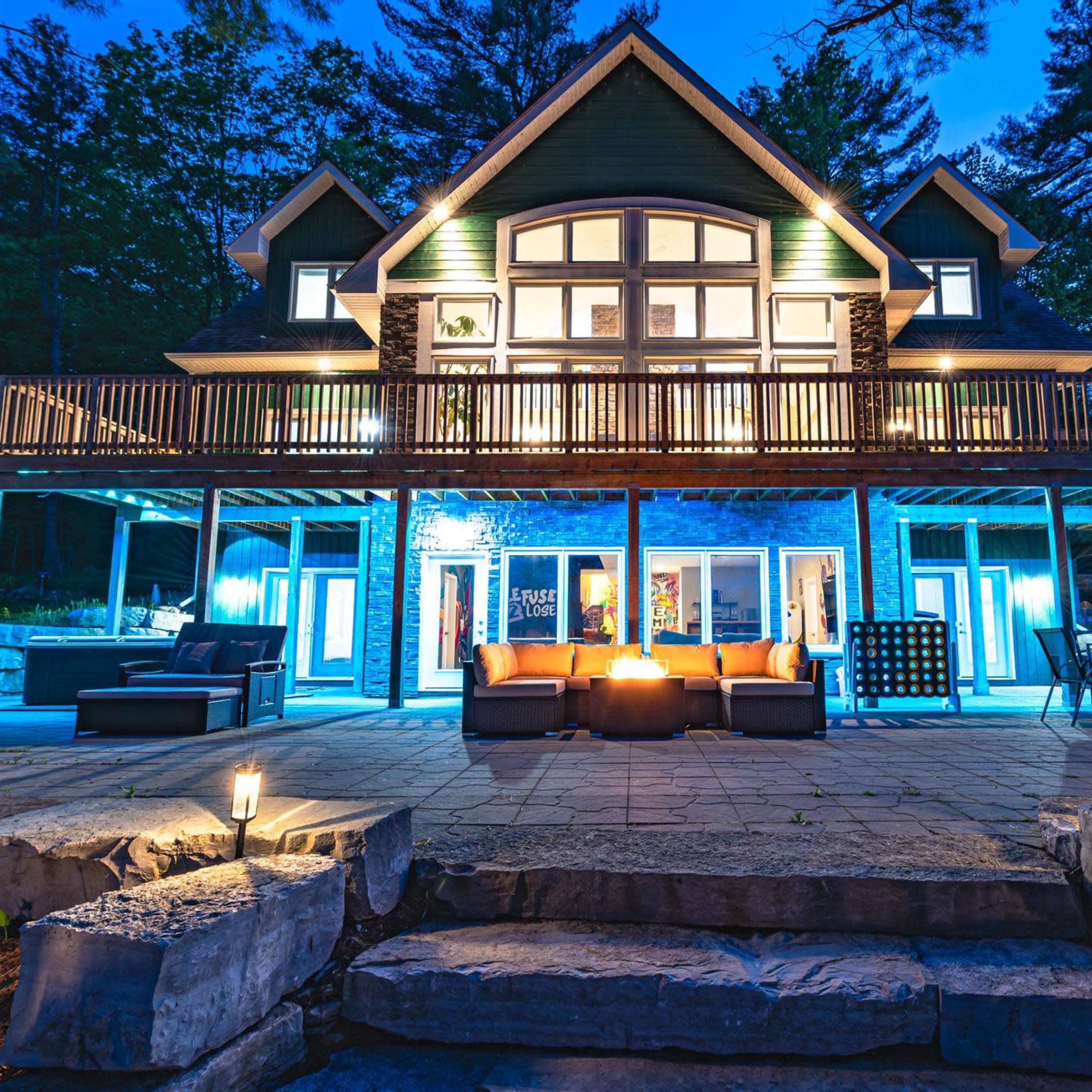 A large, modern house with a spacious deck is illuminated at dusk, featuring outdoor seating and a stone patio surrounded by trees.