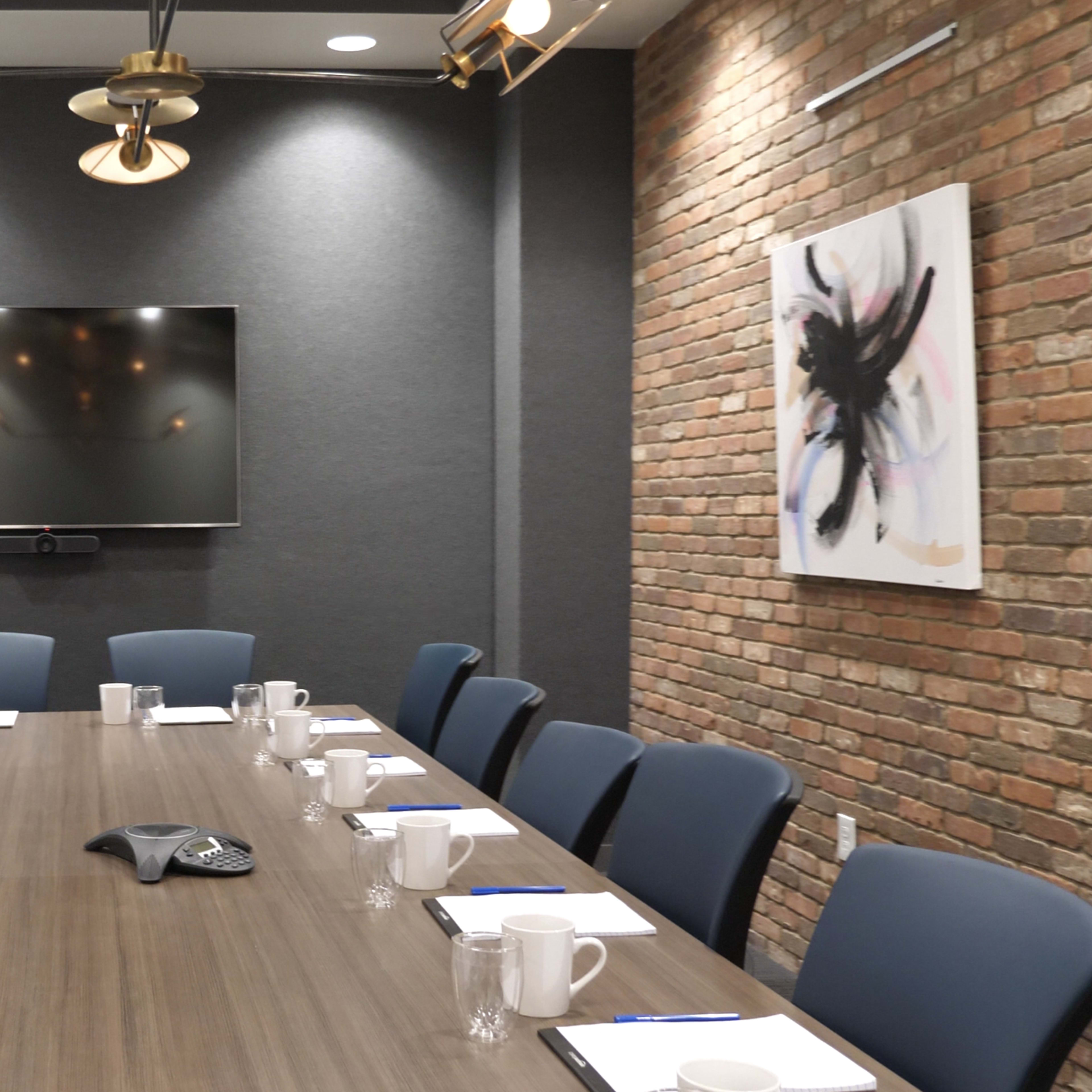 A modern conference room features a long wooden table with chairs, a large wall-mounted screen, a whiteboard, and brick accents.