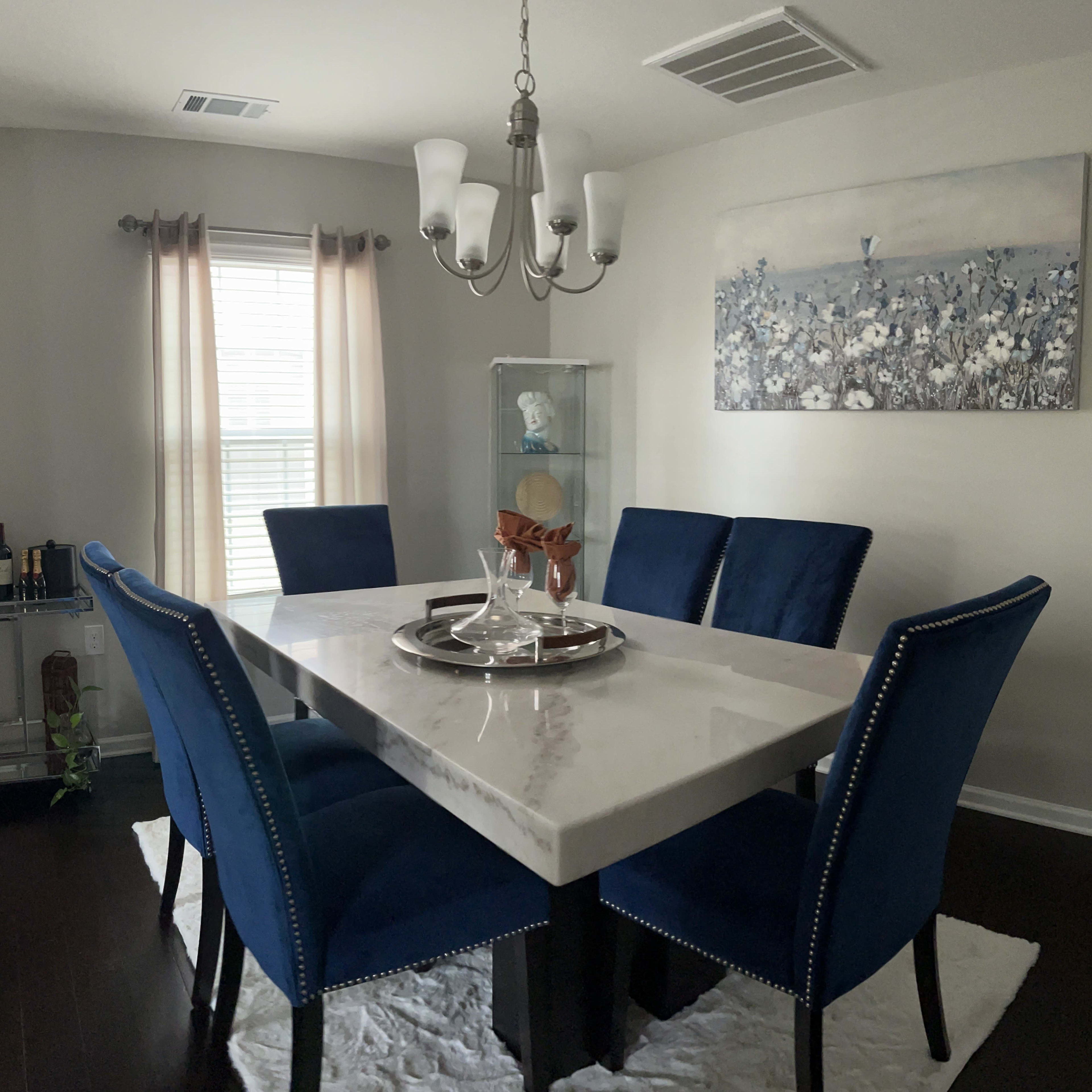 A modern dining room features a marble-top table surrounded by four blue upholstered chairs, with a chandelier overhead and a flower-themed artwork on the wall.