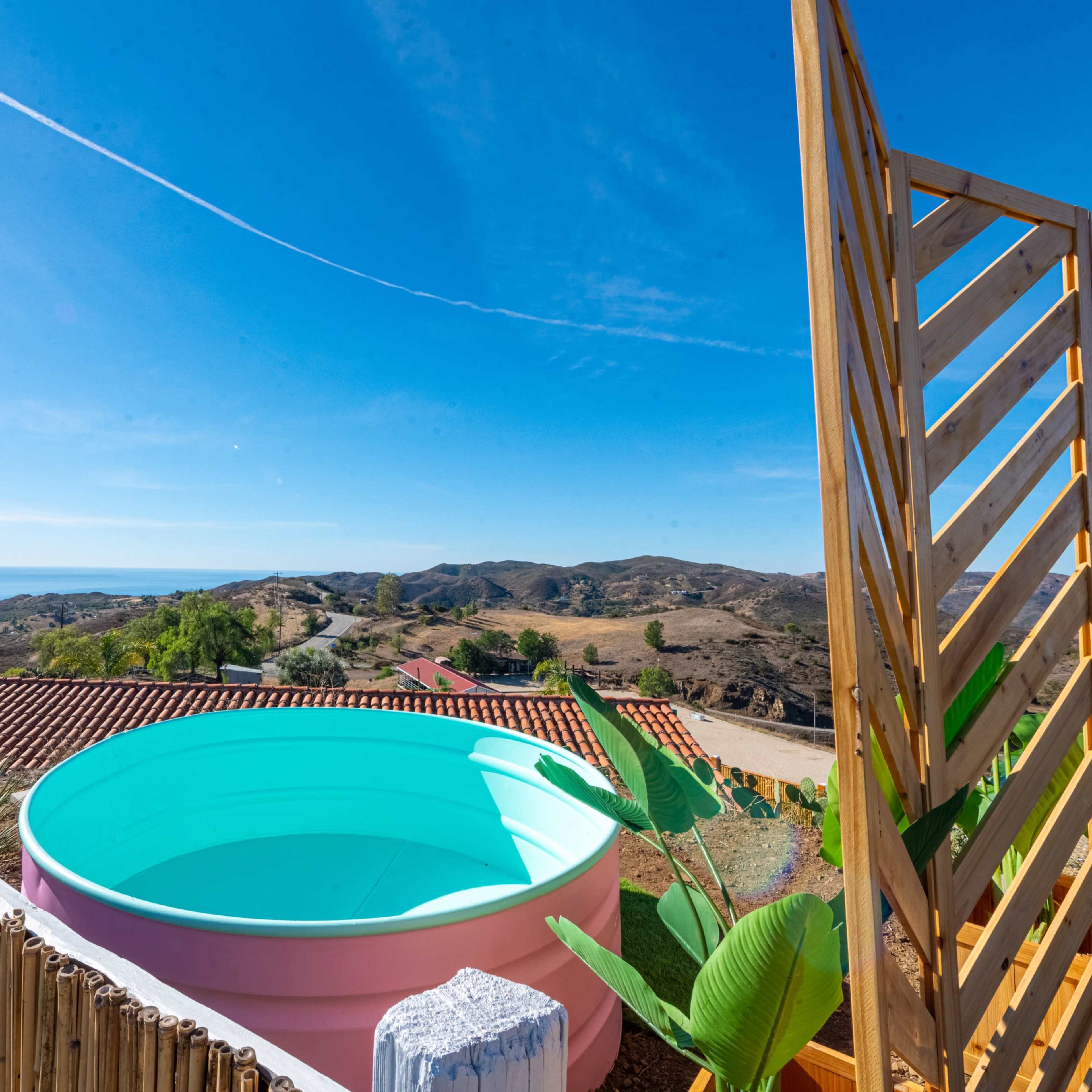 A blue circular pool is situated on a hillside overlooking a landscape of rolling hills and distant ocean views under a clear blue sky.