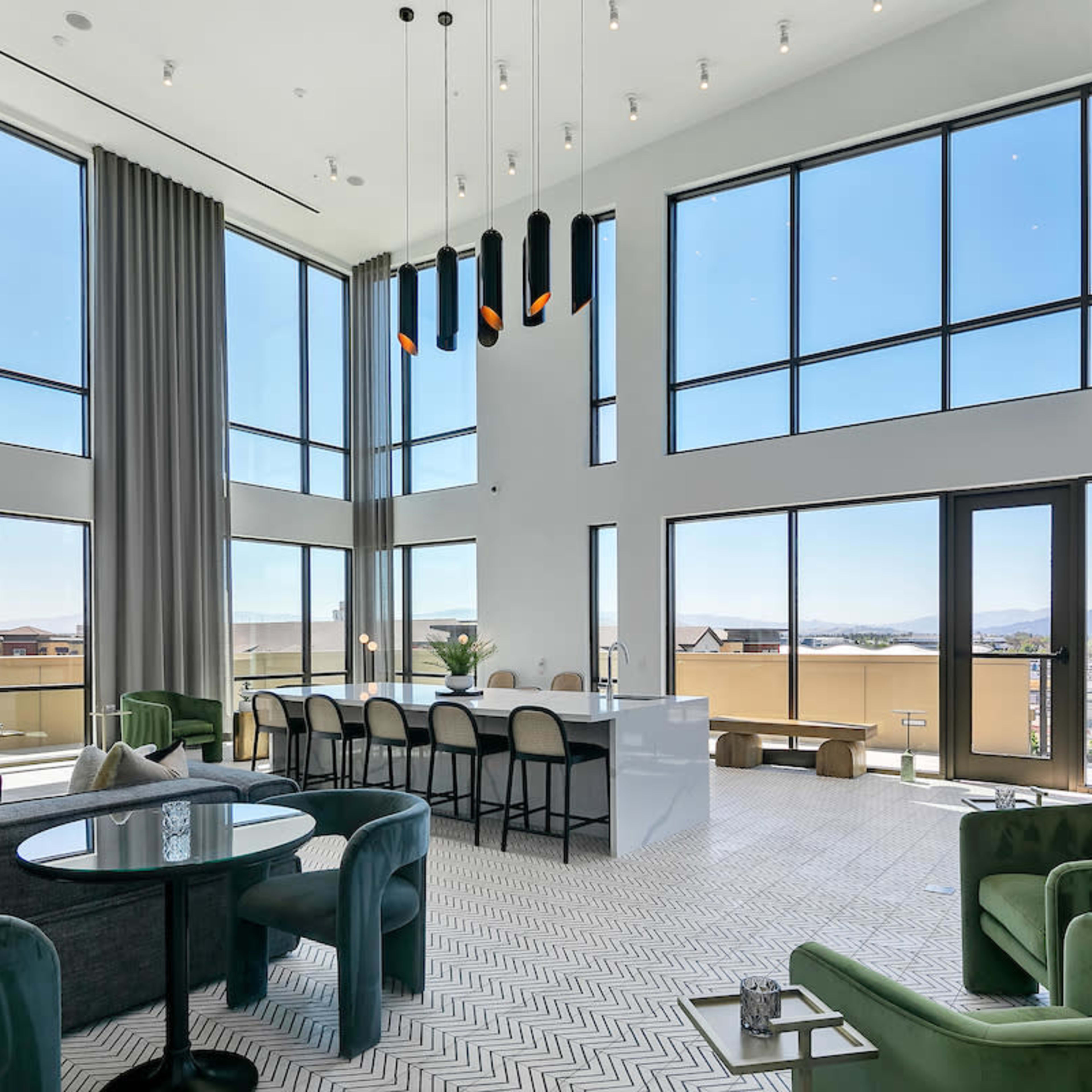 A modern living space with large windows, high ceilings, and a kitchen area featuring a white countertop and bar seating.