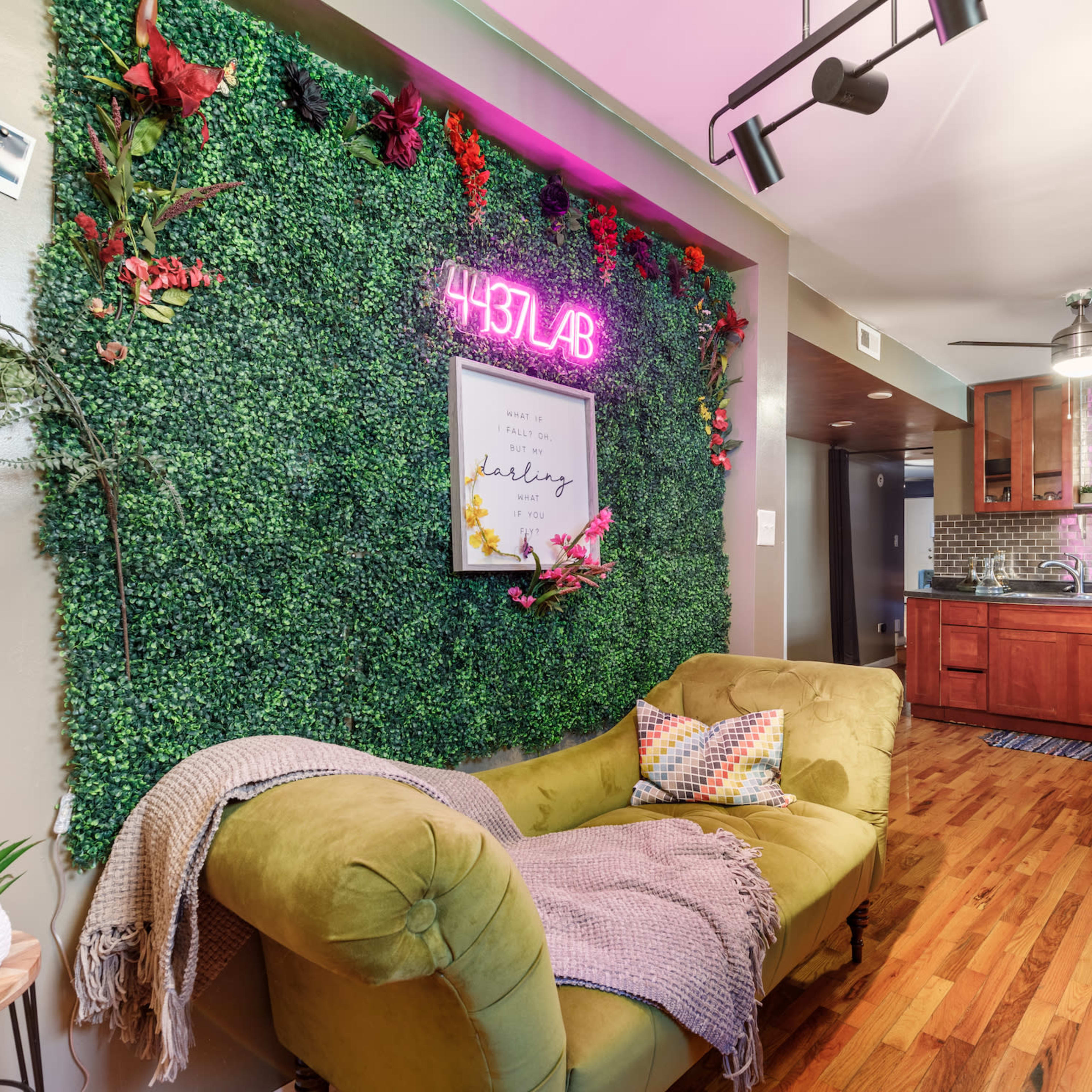 A cozy living space featuring a green wall of faux foliage with a neon sign, a yellow couch with a throw blanket and pillows, and a kitchen area in the background with wooden cabinetry.