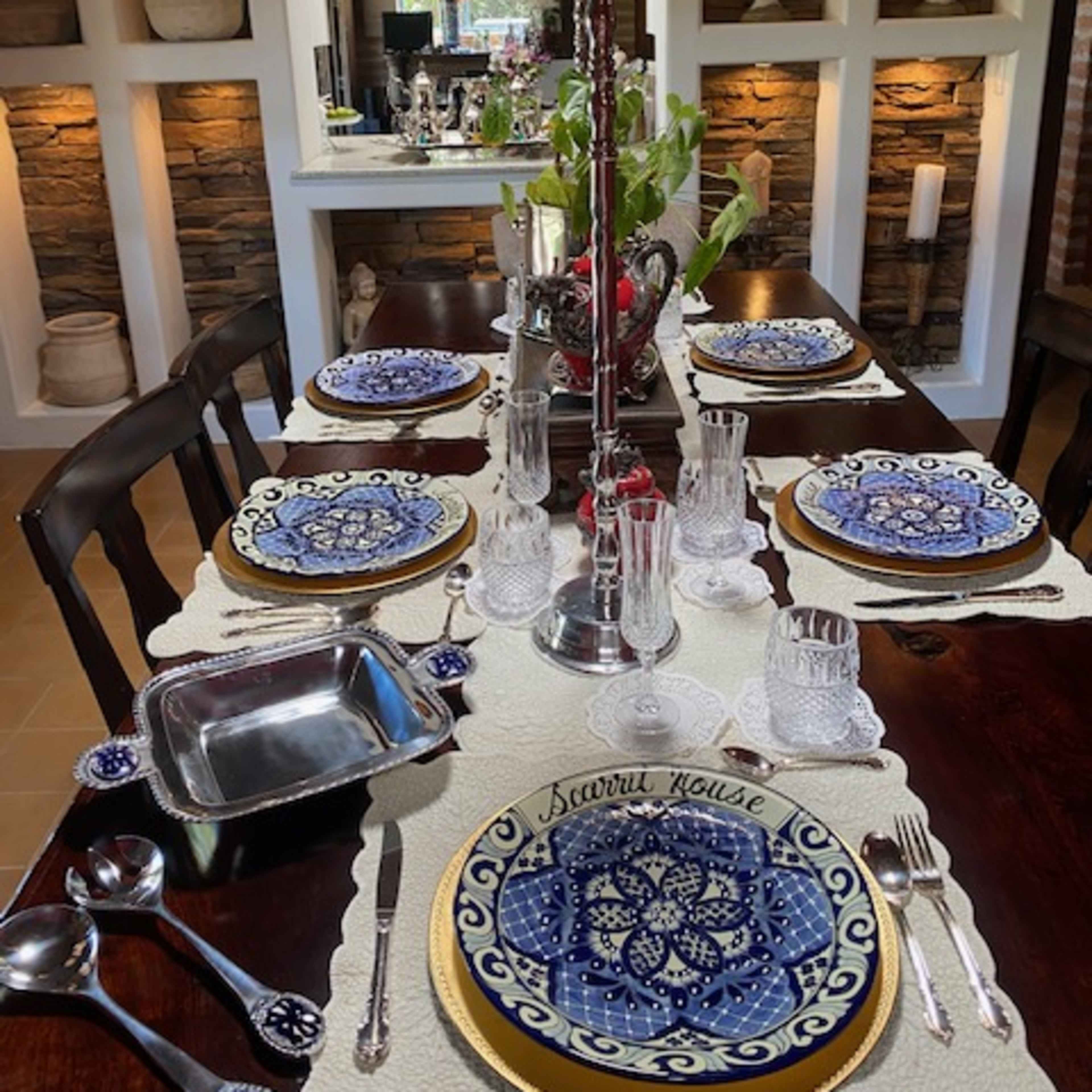 A dining table is set with blue and white patterned plates, glassware, and a centerpiece candle holder, surrounded by stone wall accents in the background.