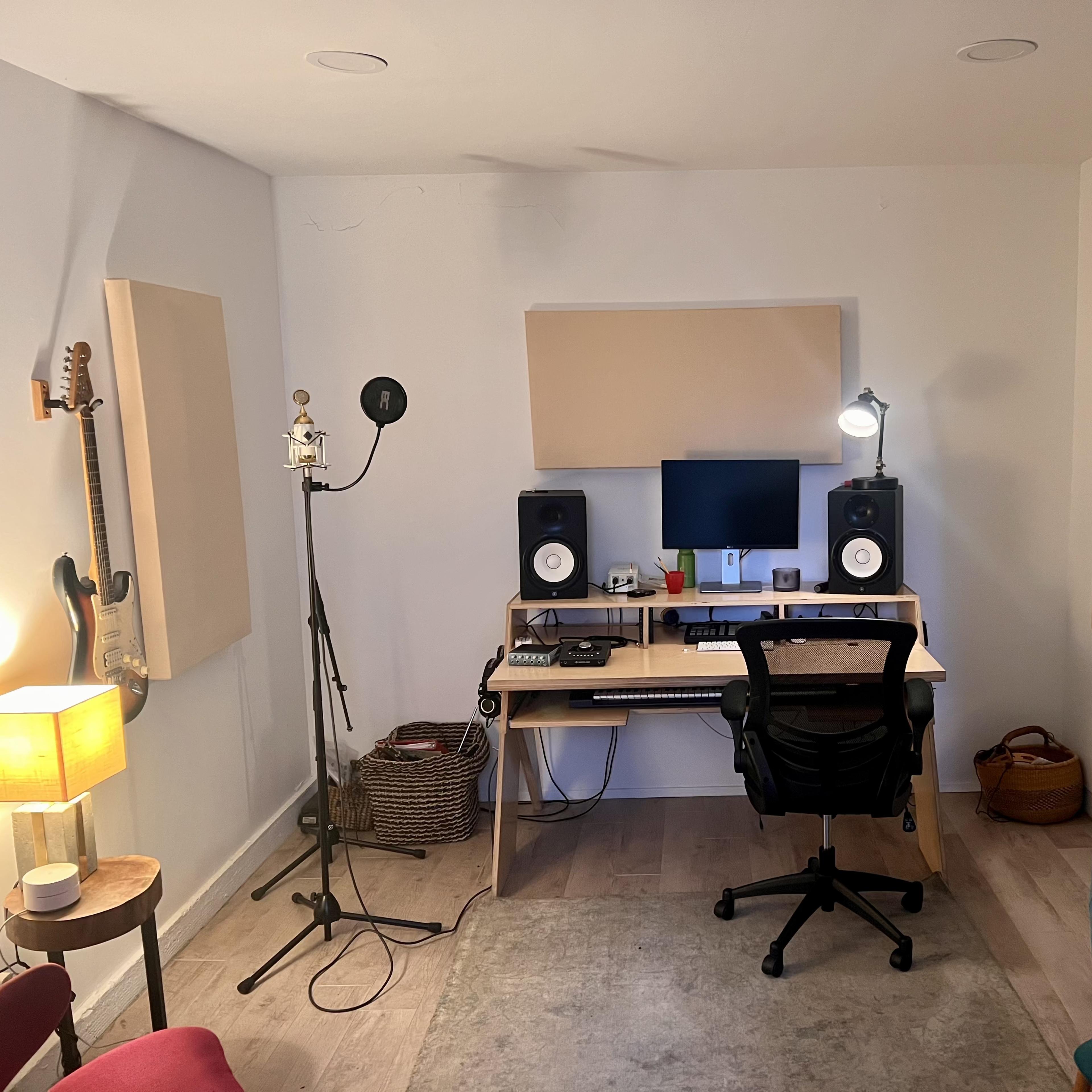 A small music studio with a desk, computer, speakers, a microphone on a stand, and guitars mounted on the walls.