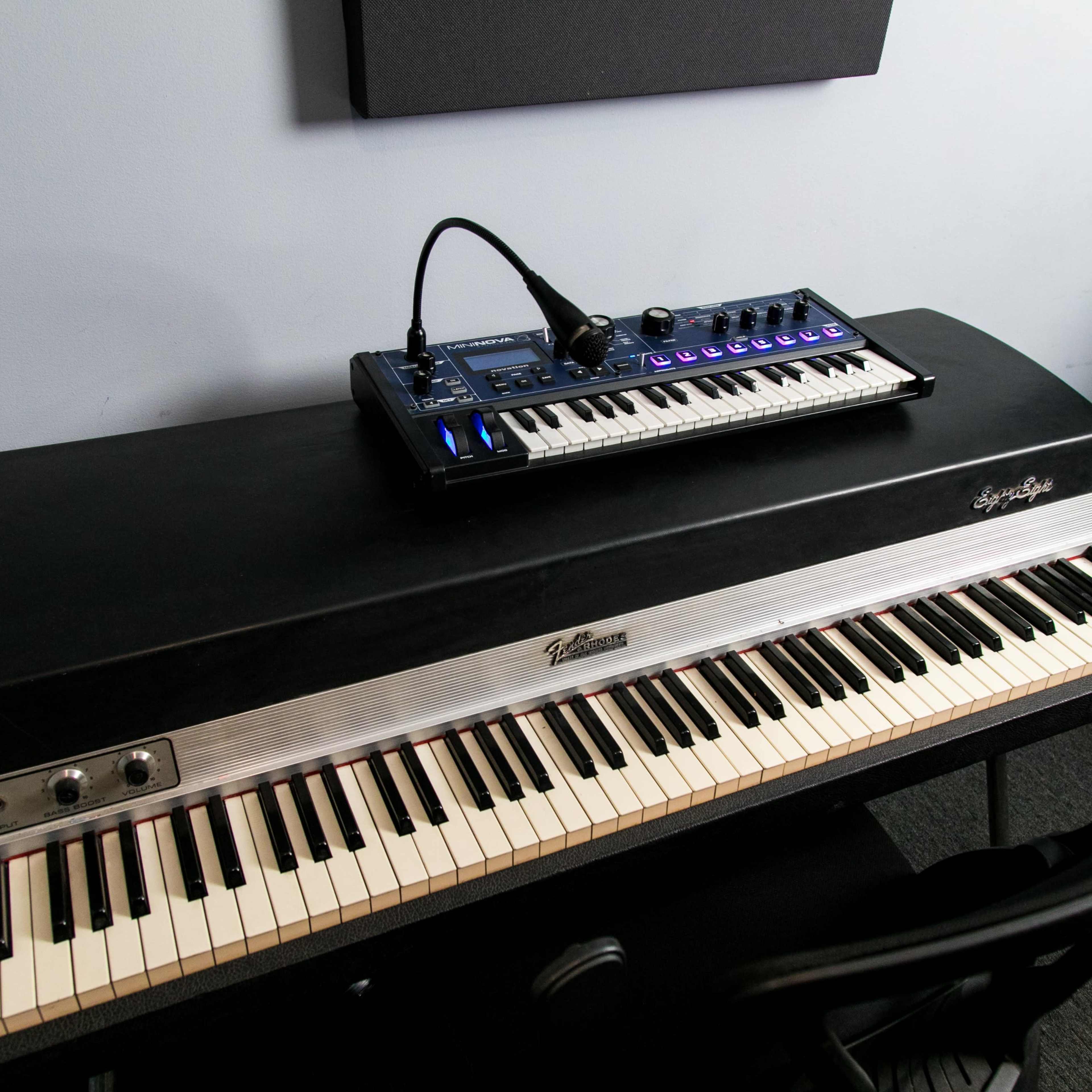 A Rhodes electric piano and a synthesizer are placed on a black keyboard stand in a music studio setting.