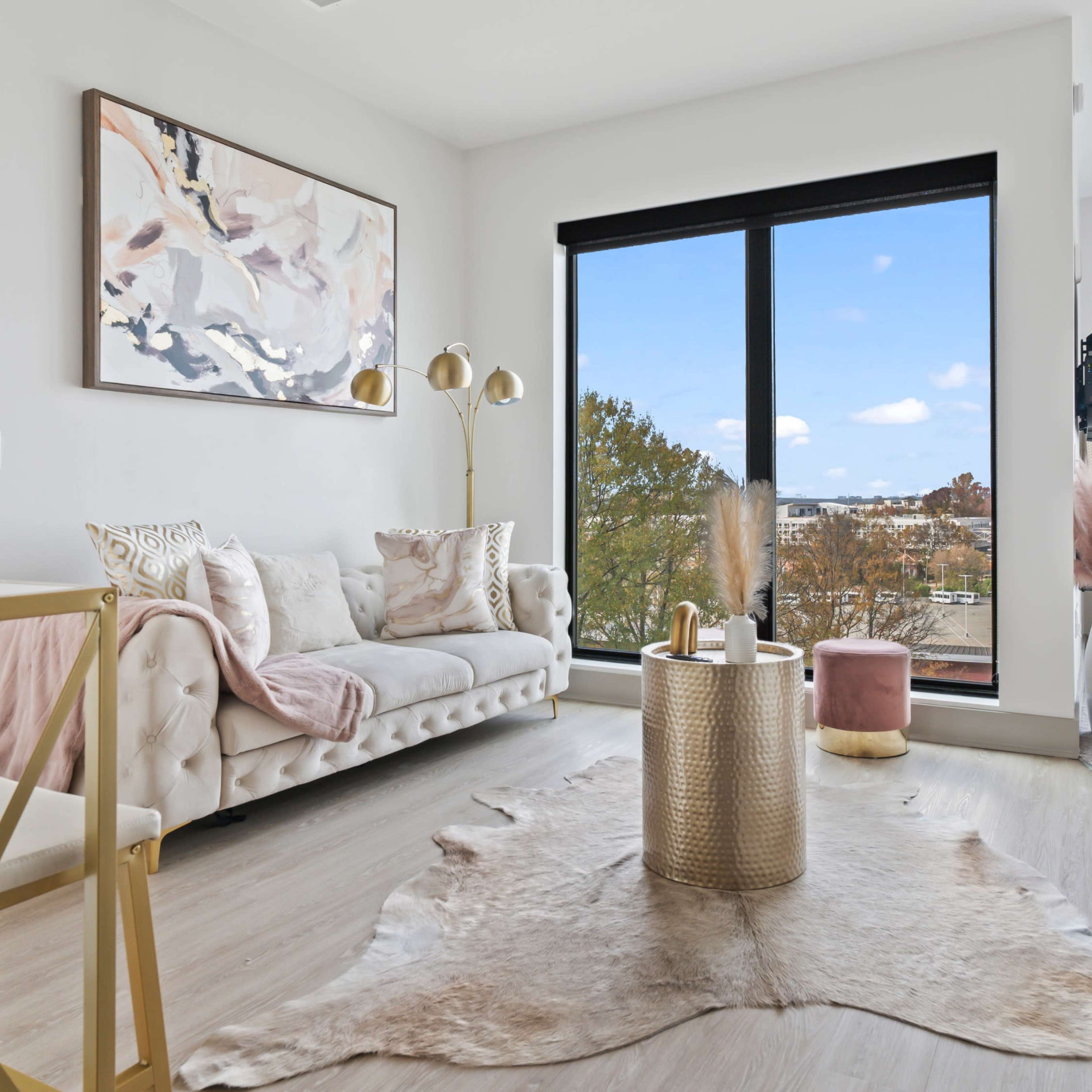 The scene features a modern living room with a light-colored sofa, a round gold coffee table, a decorative rug, and large windows showing an outdoor view.