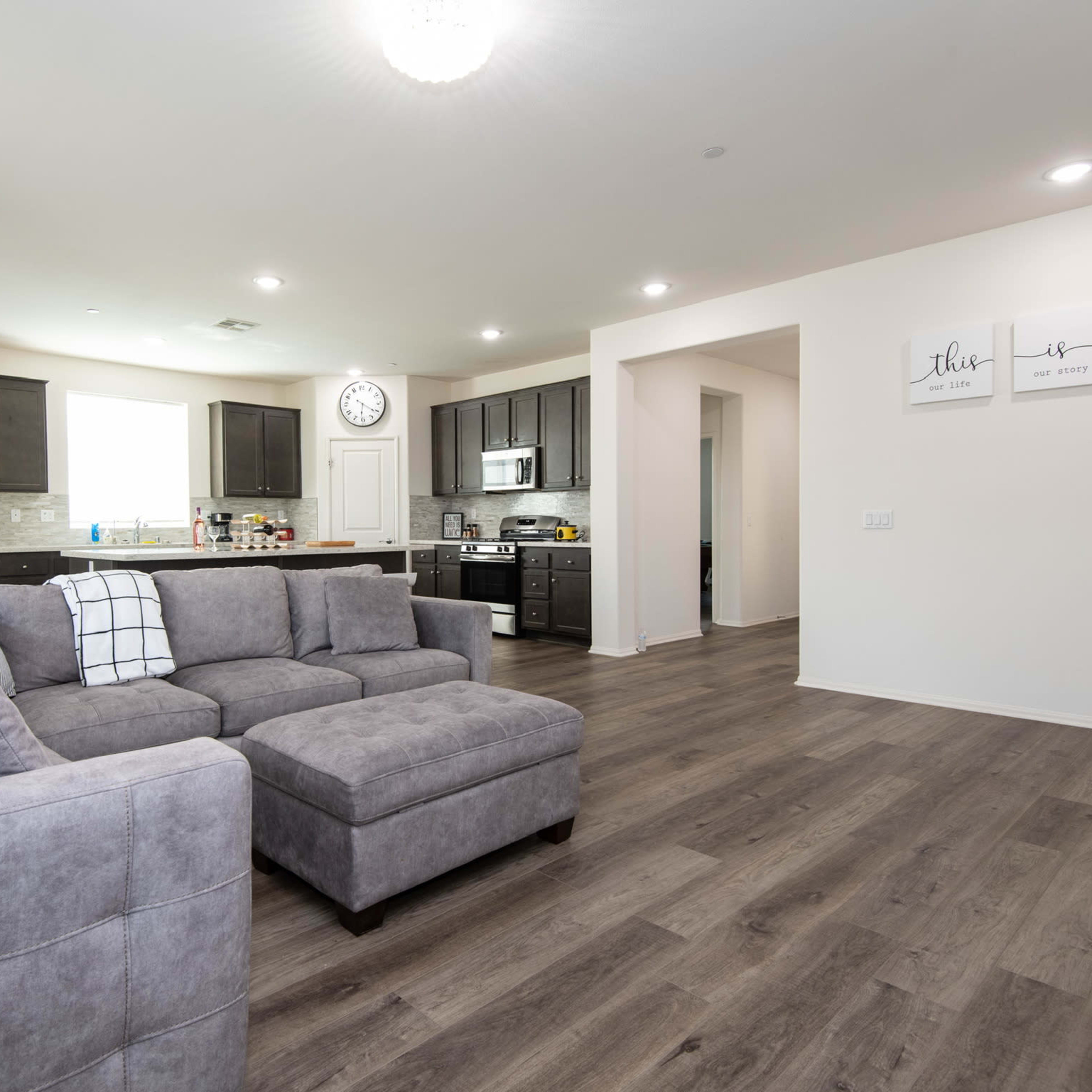 A modern living room features a gray sectional sofa, hardwood floors, and an open kitchen with dark cabinets and stainless steel appliances.