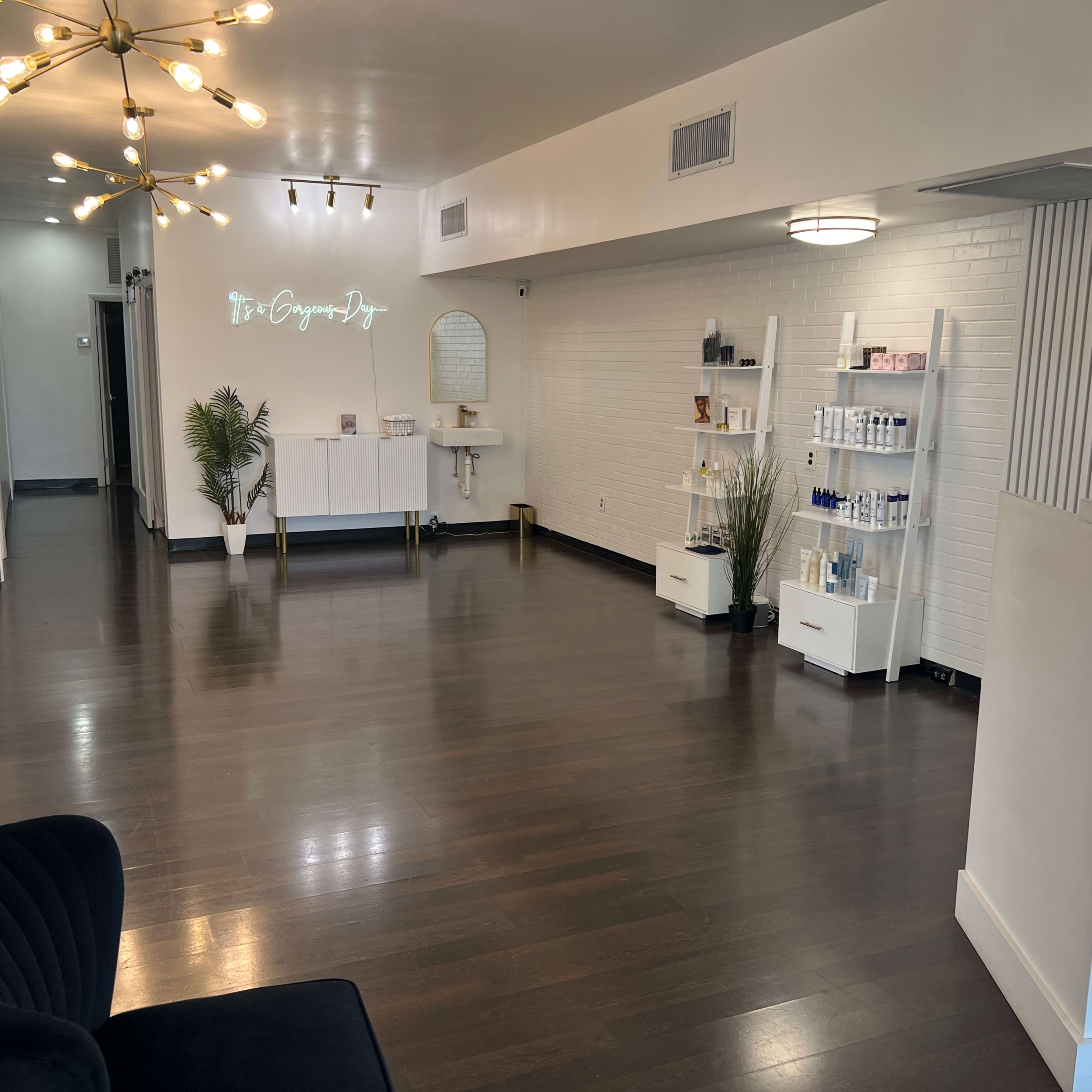 A modern, spacious reception area of a beauty or wellness clinic, featuring a central lighting fixture, white walls, and shelves displaying various products.