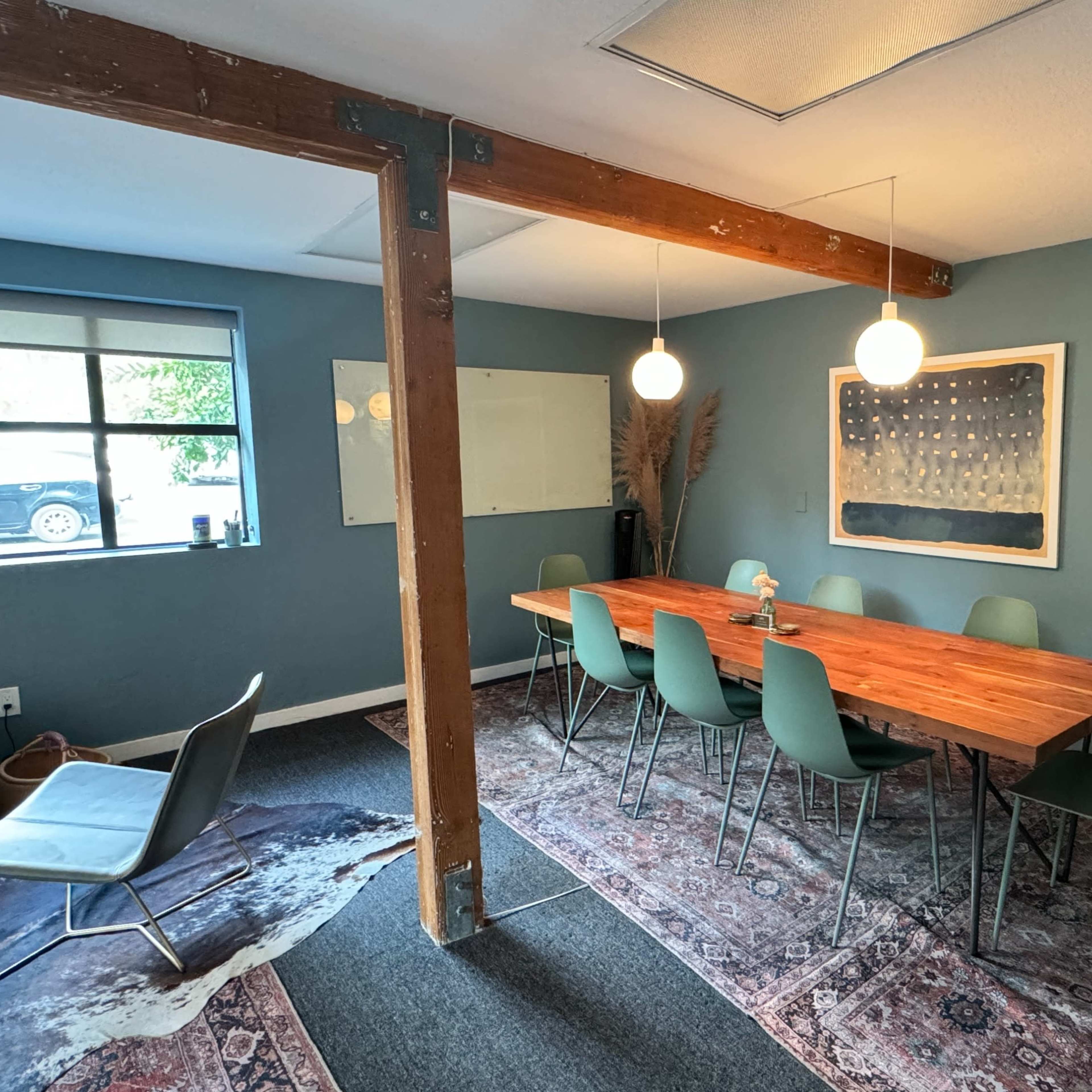 A conference room features a long wooden table surrounded by green chairs, with a large wall art piece, a whiteboard, and a window showing a parked car outside.