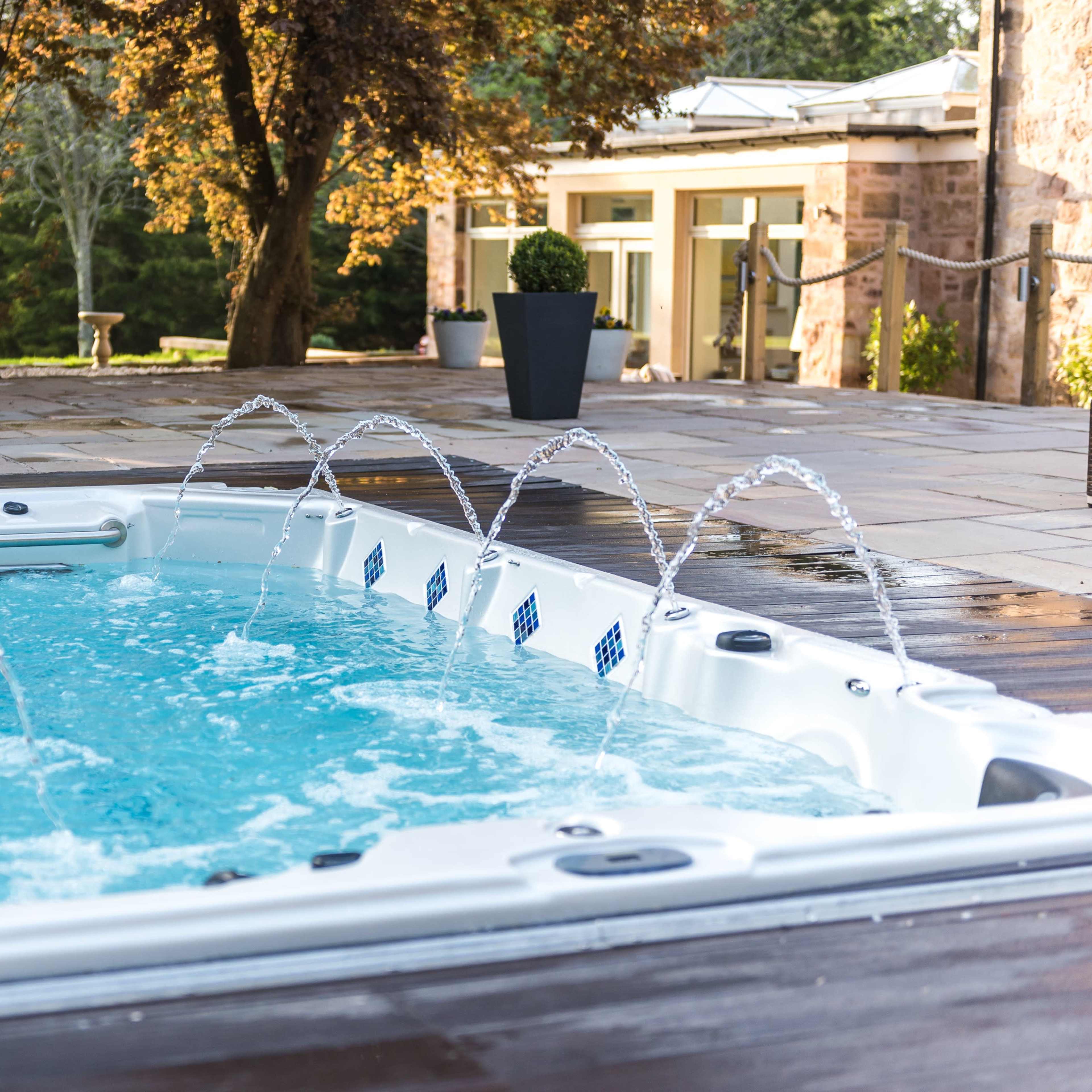 A hot tub with water jets is set on a patio surrounded by trees and a modern home.