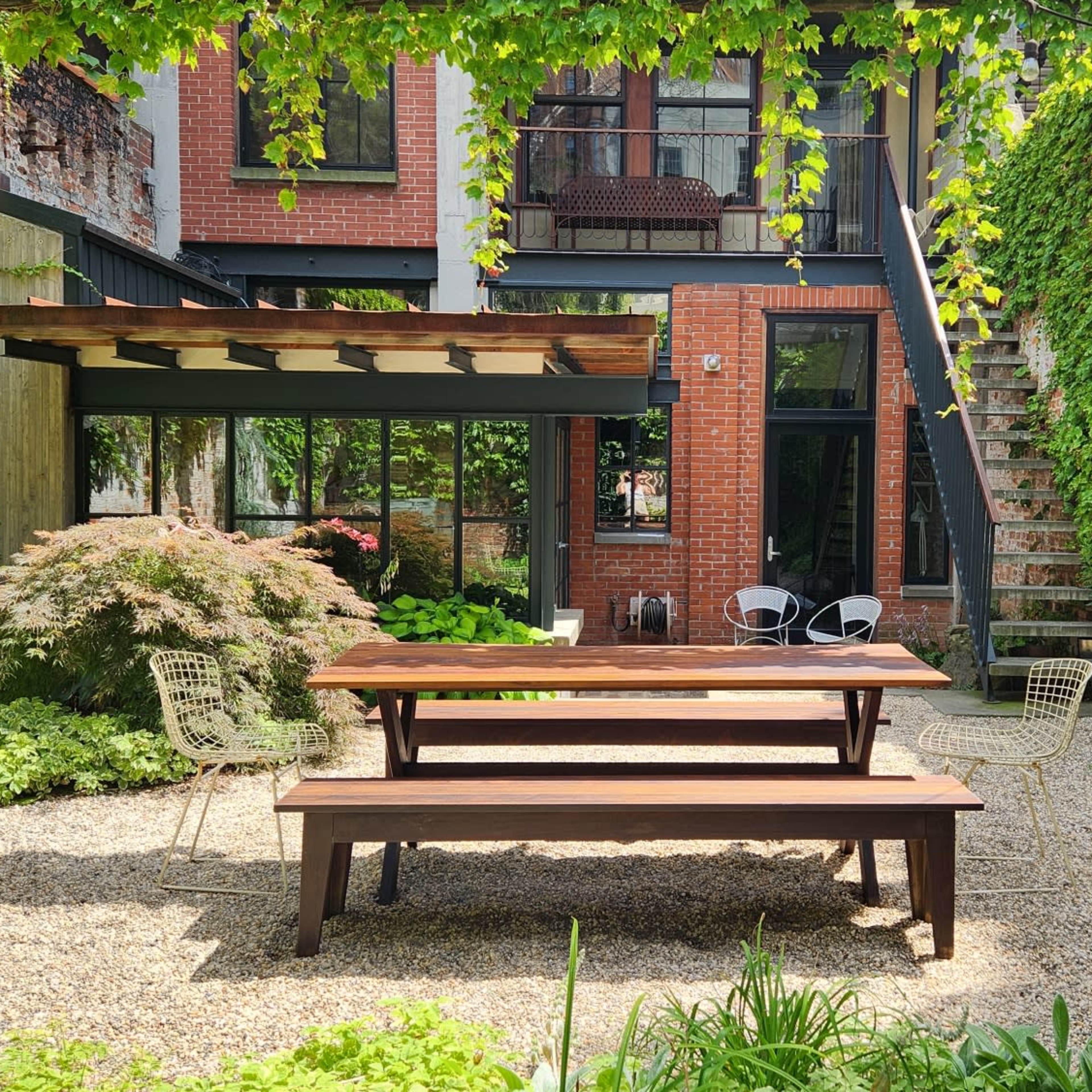 A wooden picnic table is set in a gravel courtyard surrounded by brick walls and greenery, with a staircase leading to an upper level in the background.