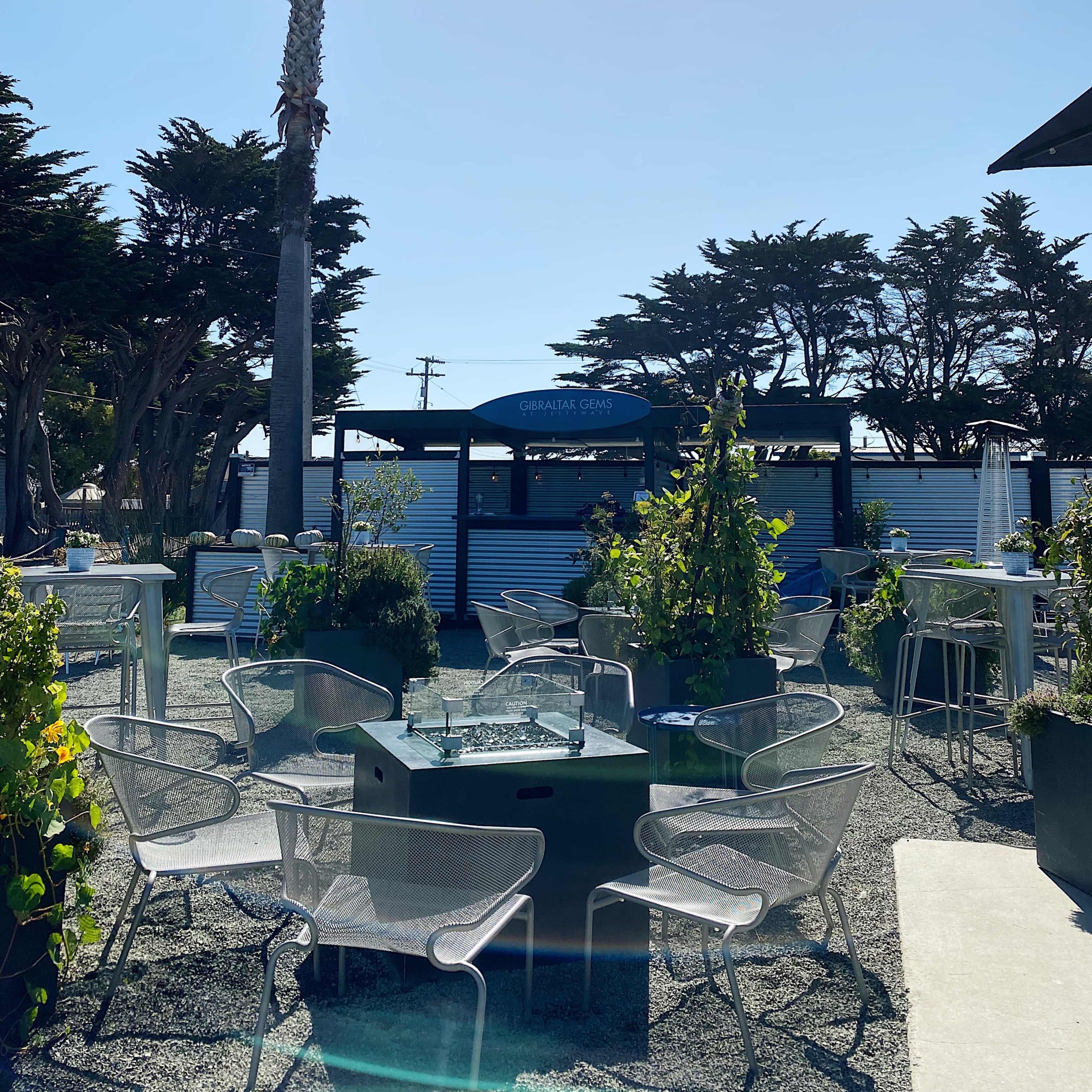 A sunny outdoor dining area features metal chairs and tables surrounded by greenery and palm trees.
