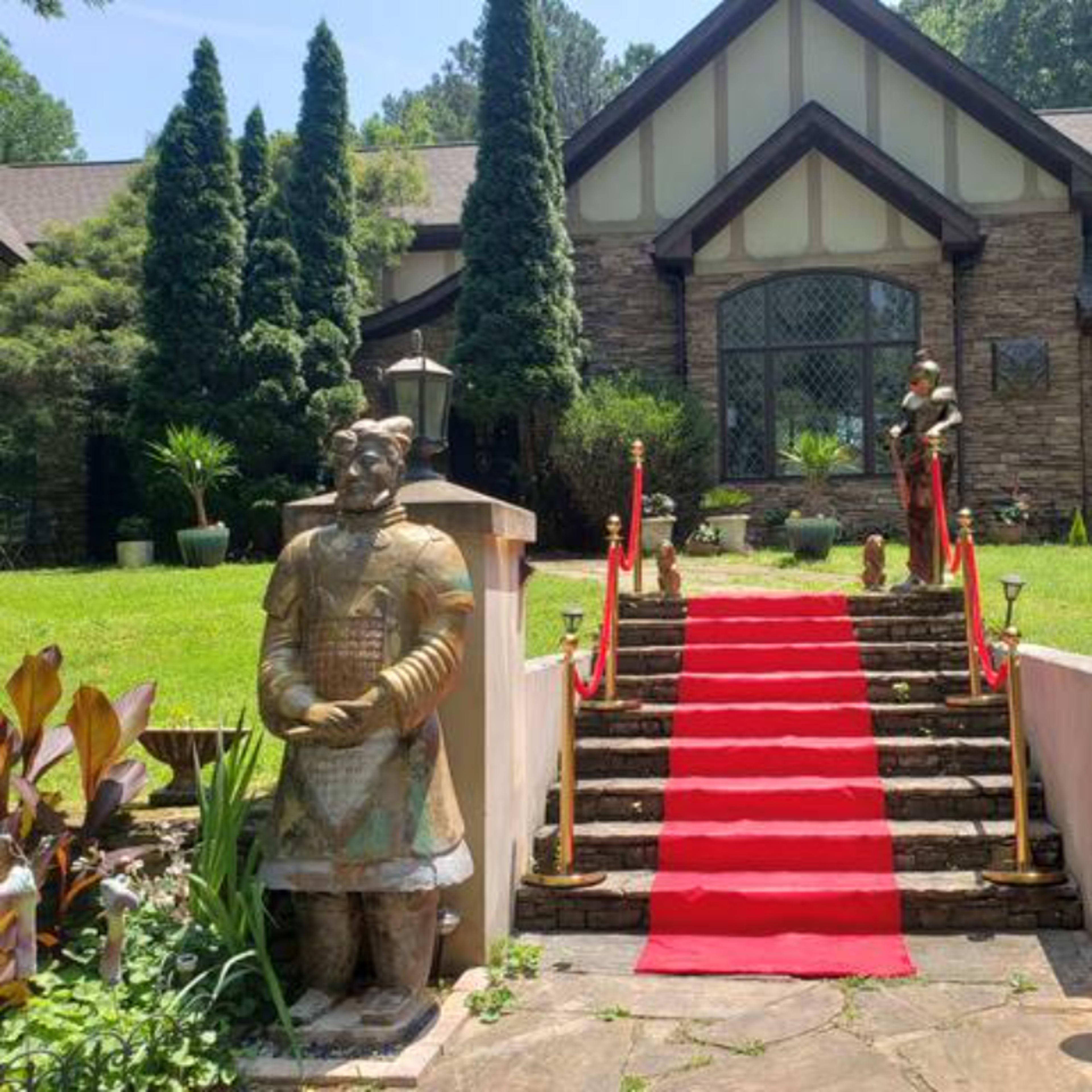 A pathway with a red carpet and two stone statues leads to the entrance of a large, stone-faced house surrounded by greenery.