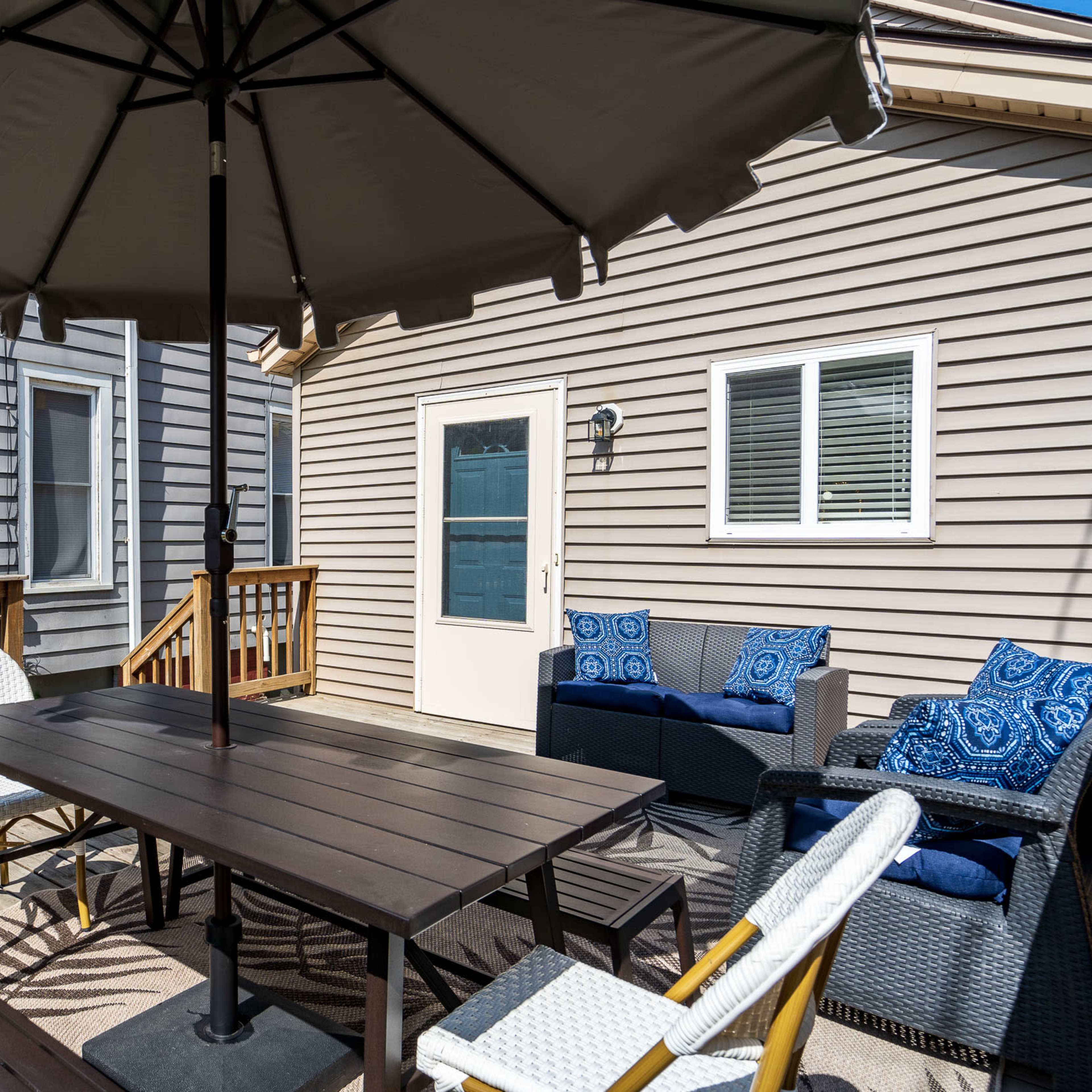 A patio area featuring a black wooden picnic table, an umbrella, a gas grill, and seating arranged against beige siding and a door with a small window.