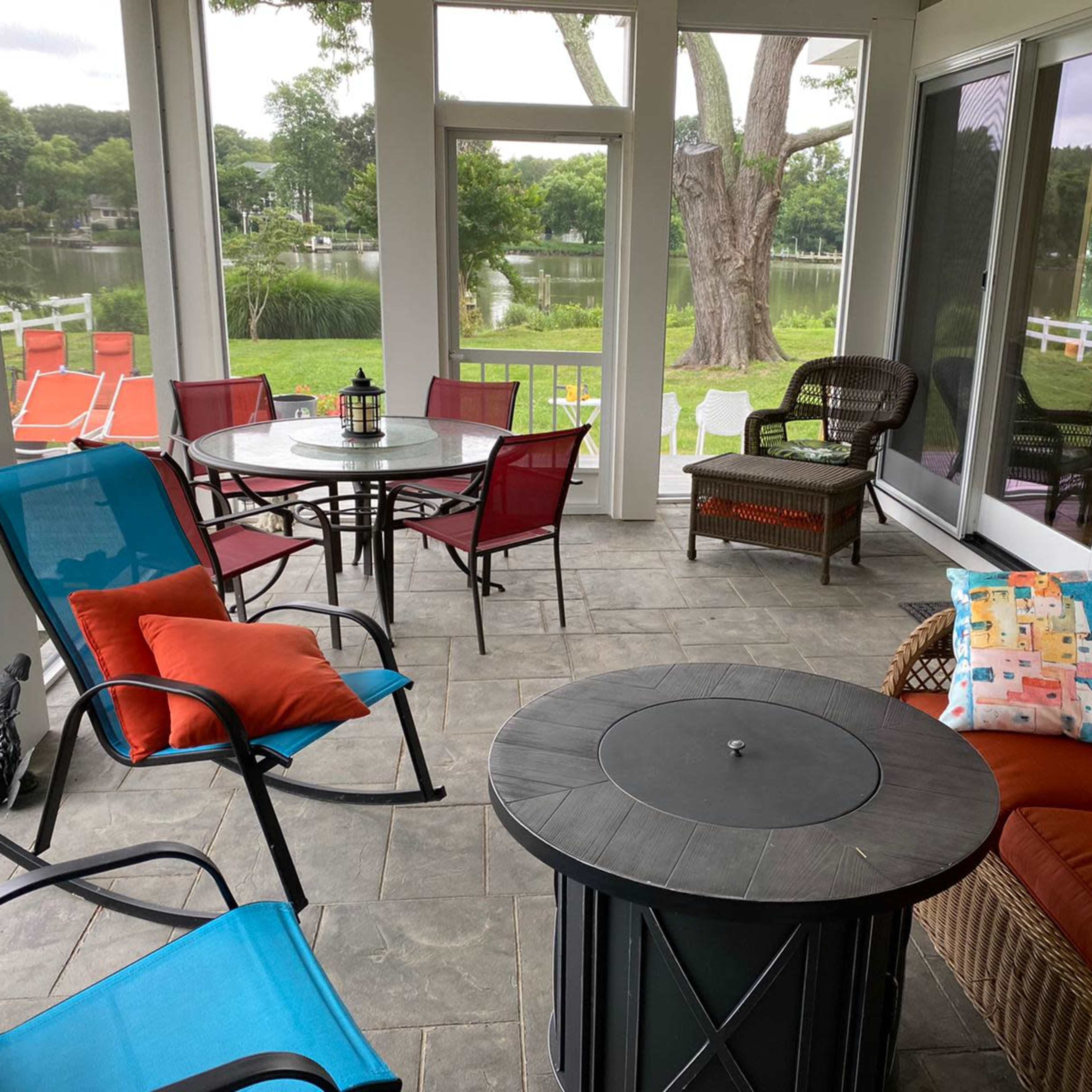 A screened porch features a round table surrounded by colorful chairs and a cozy seating area, overlooking a lake.