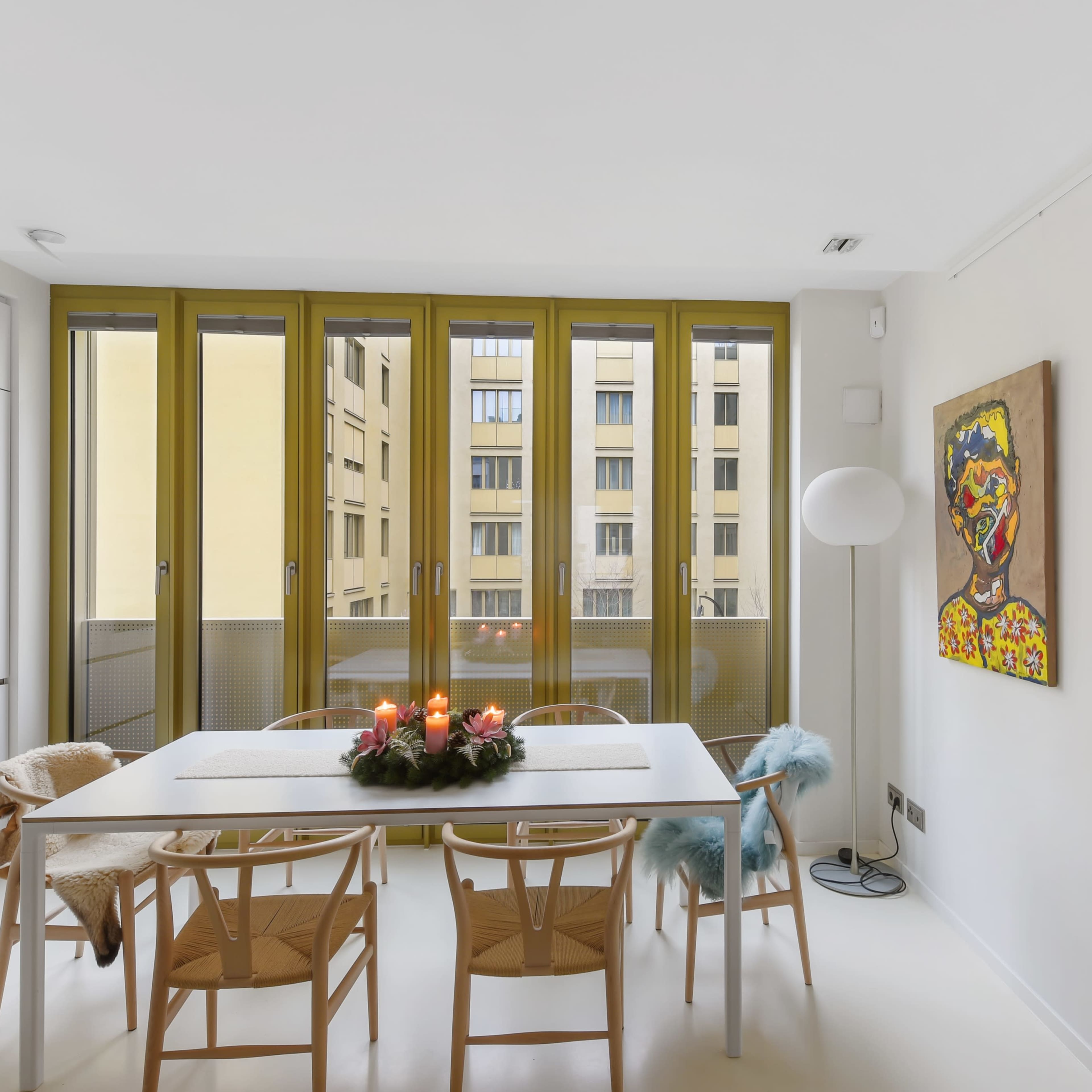 A minimalist dining area with a table set for four, featuring a centerpiece of candles, and large windows overlooking a building courtyard.