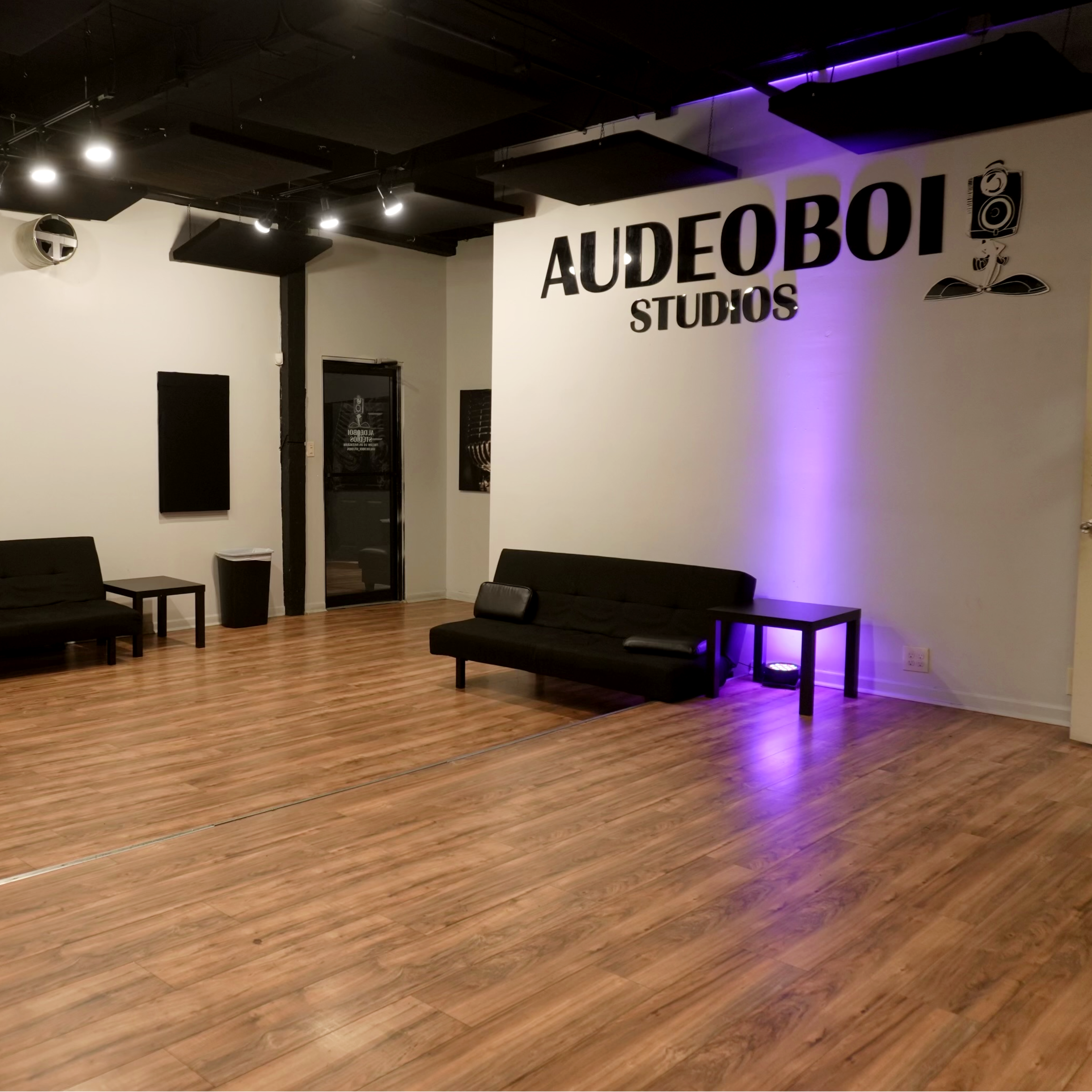 A modern studio space with black sofas, a central table, and soundproof panels on the walls, featuring the name "AUDEOBOI STUDIOS" prominently displayed.