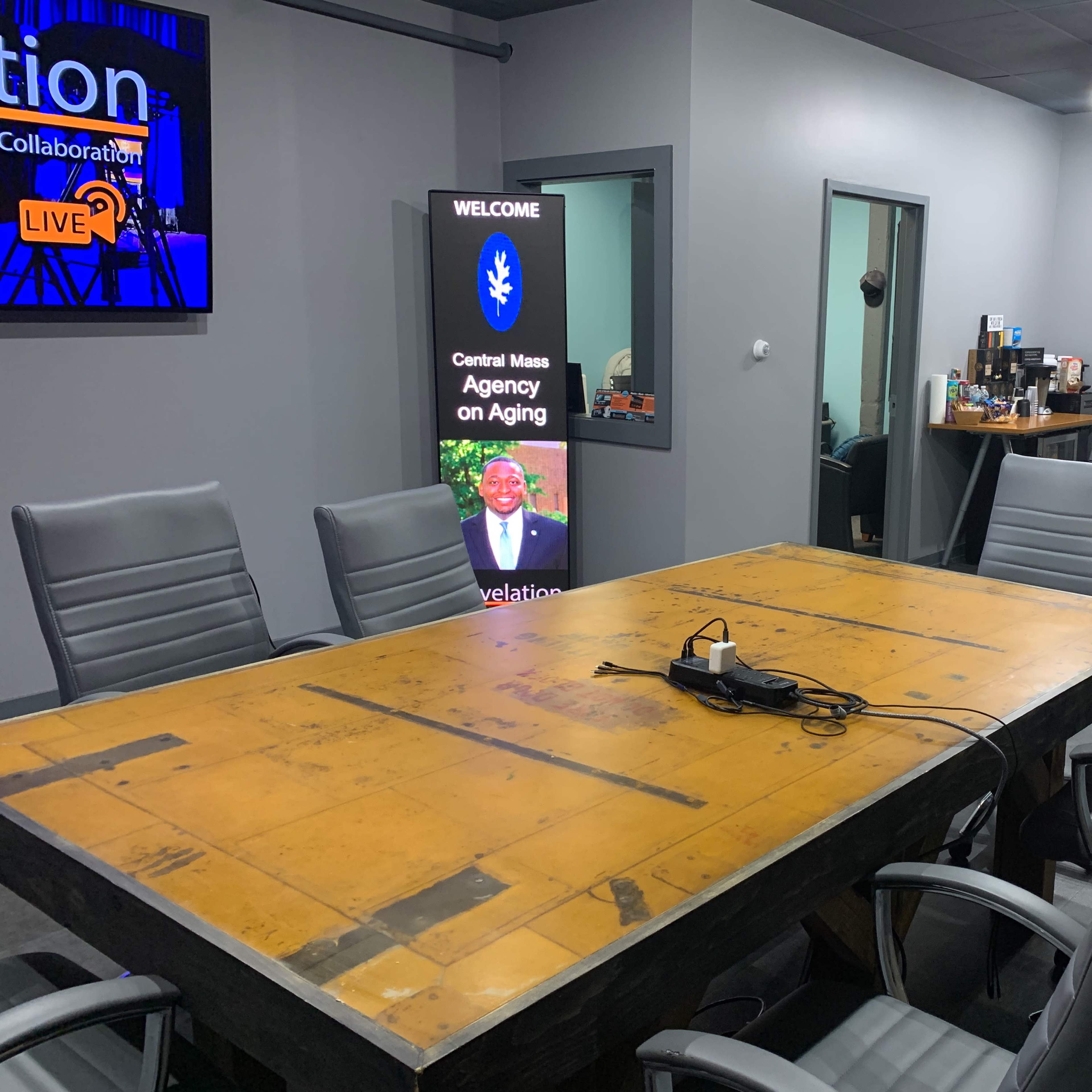 A conference room features a large wooden table surrounded by black leather chairs, with a digital display on one wall welcoming the Central Mass Agency on Aging.