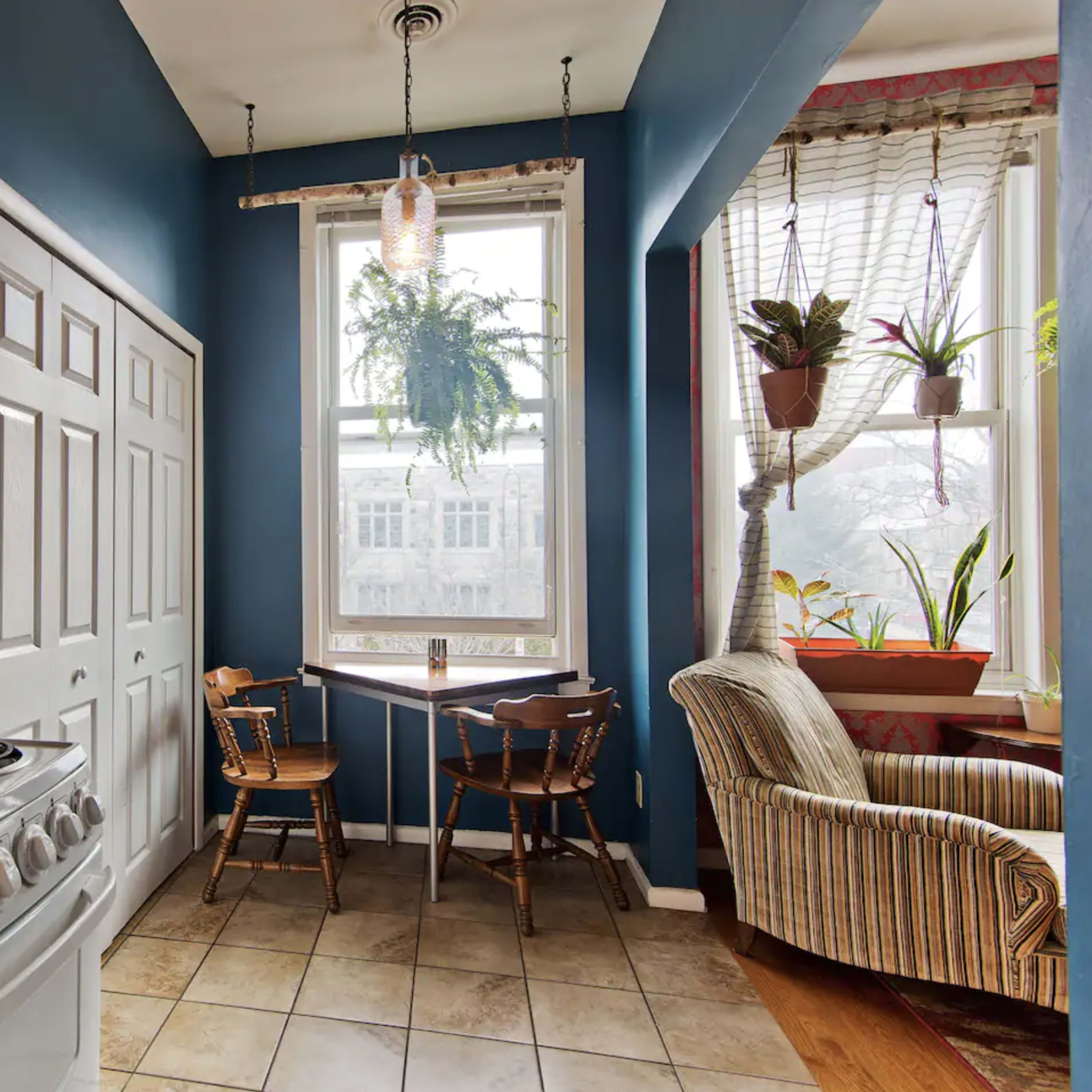 A compact kitchen features a gas stove, white cabinets, and a cozy dining nook with a table and chairs next to a large window adorned with plants.