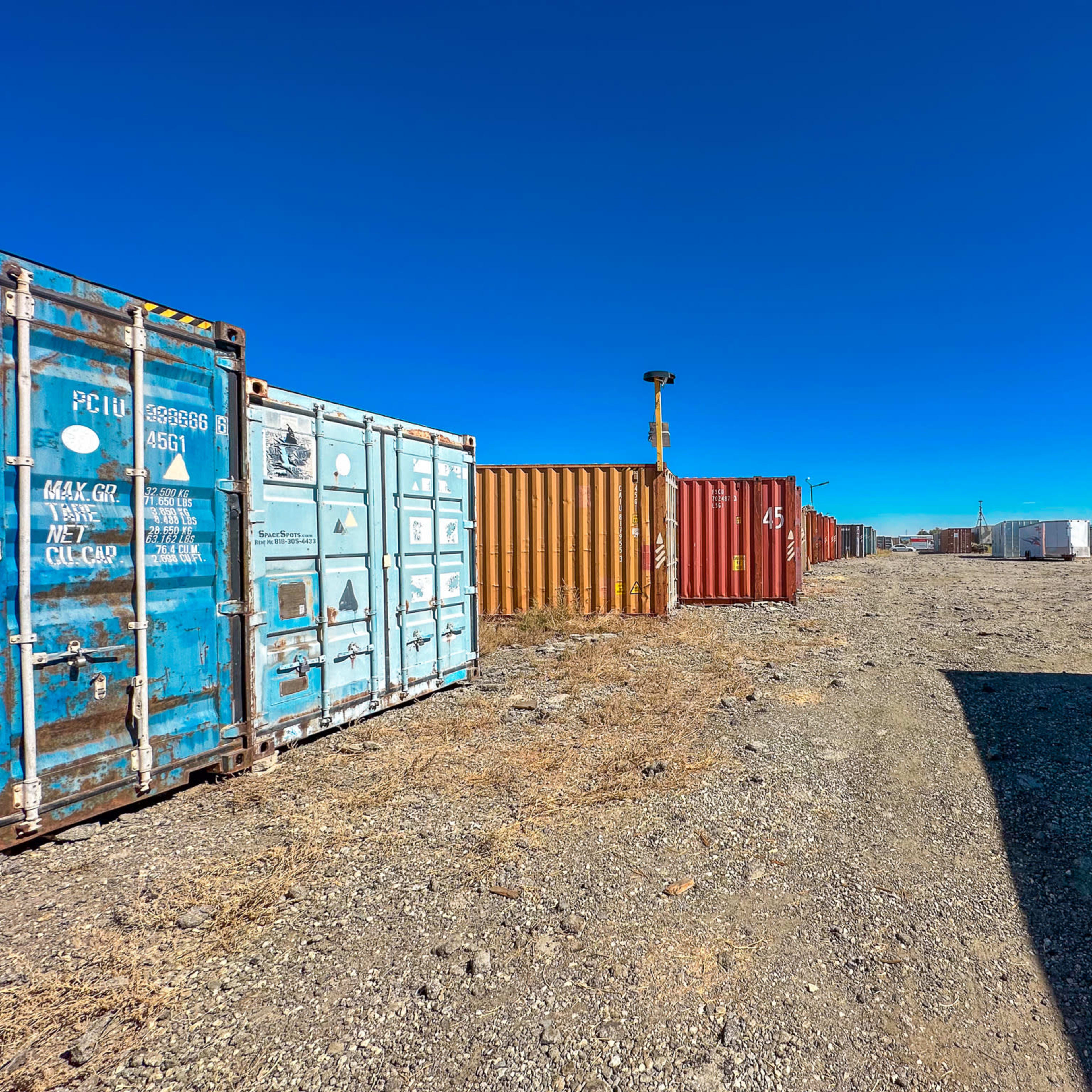 A row of shipping containers in various colors lined along a gravel path under a clear blue sky.