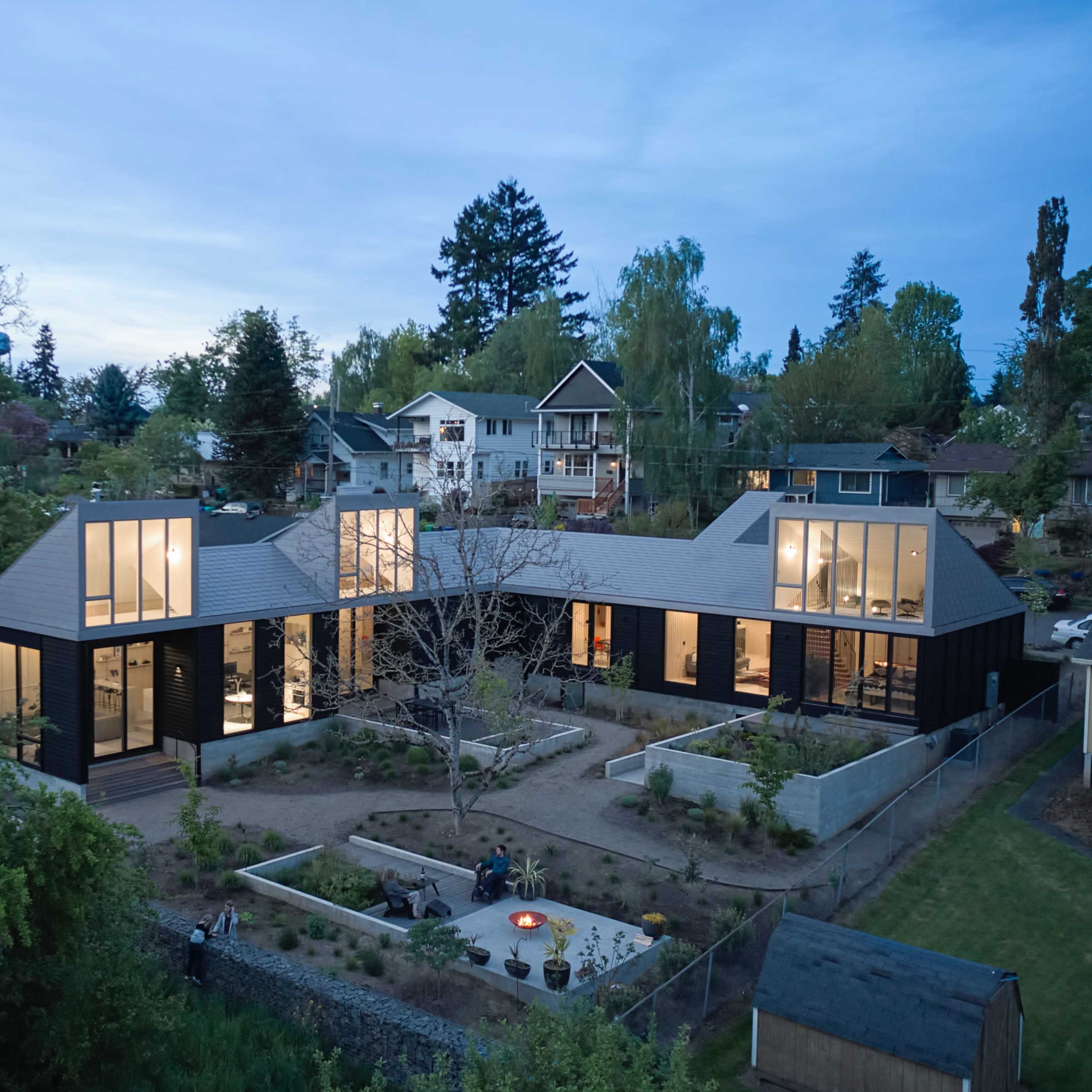 A modern house with large windows is surrounded by greenery and features an outdoor patio with a fire pit.
