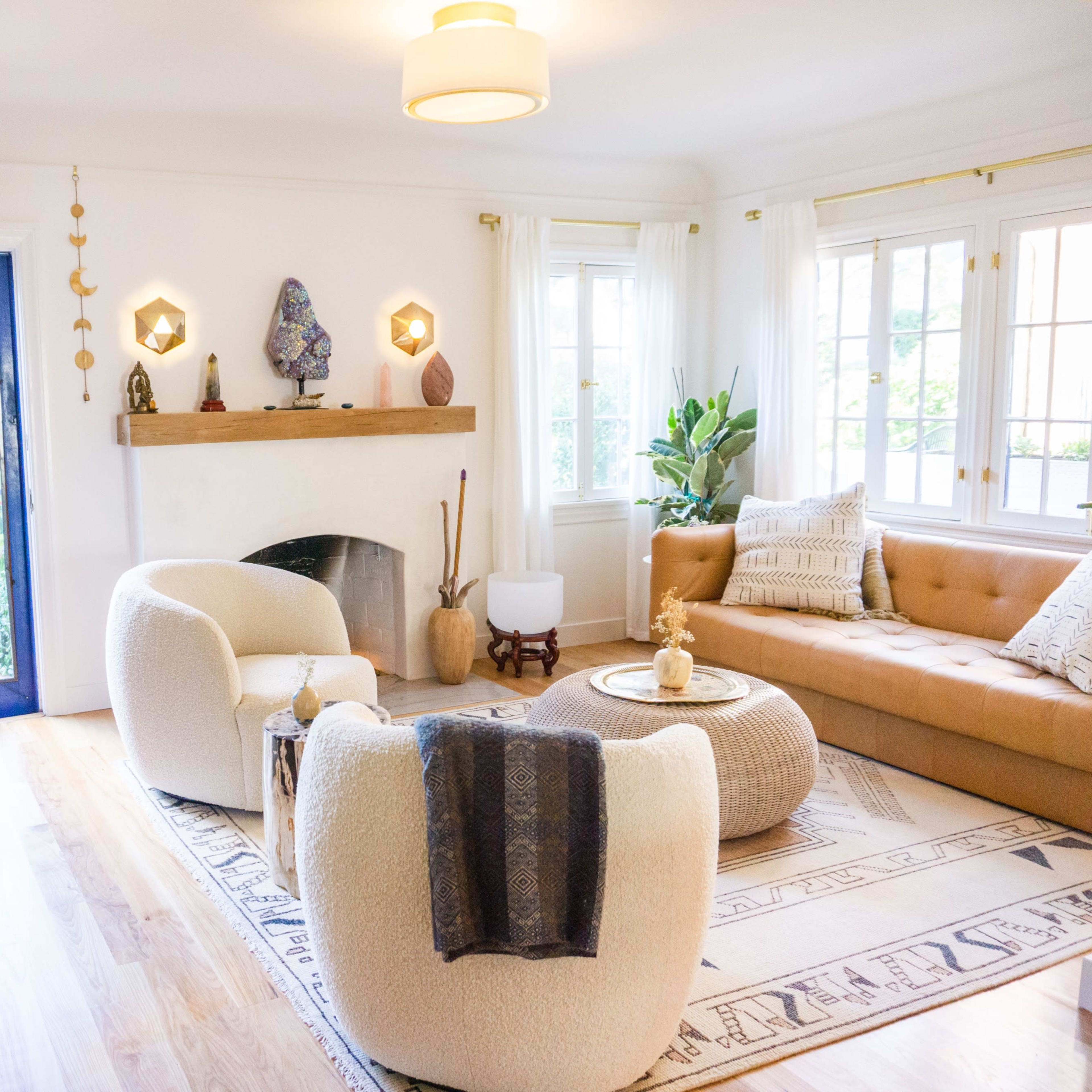 A cozy living room features a tan sofa, two white armchairs, a round coffee table, and large windows overlooking a blue door and outdoor area.
