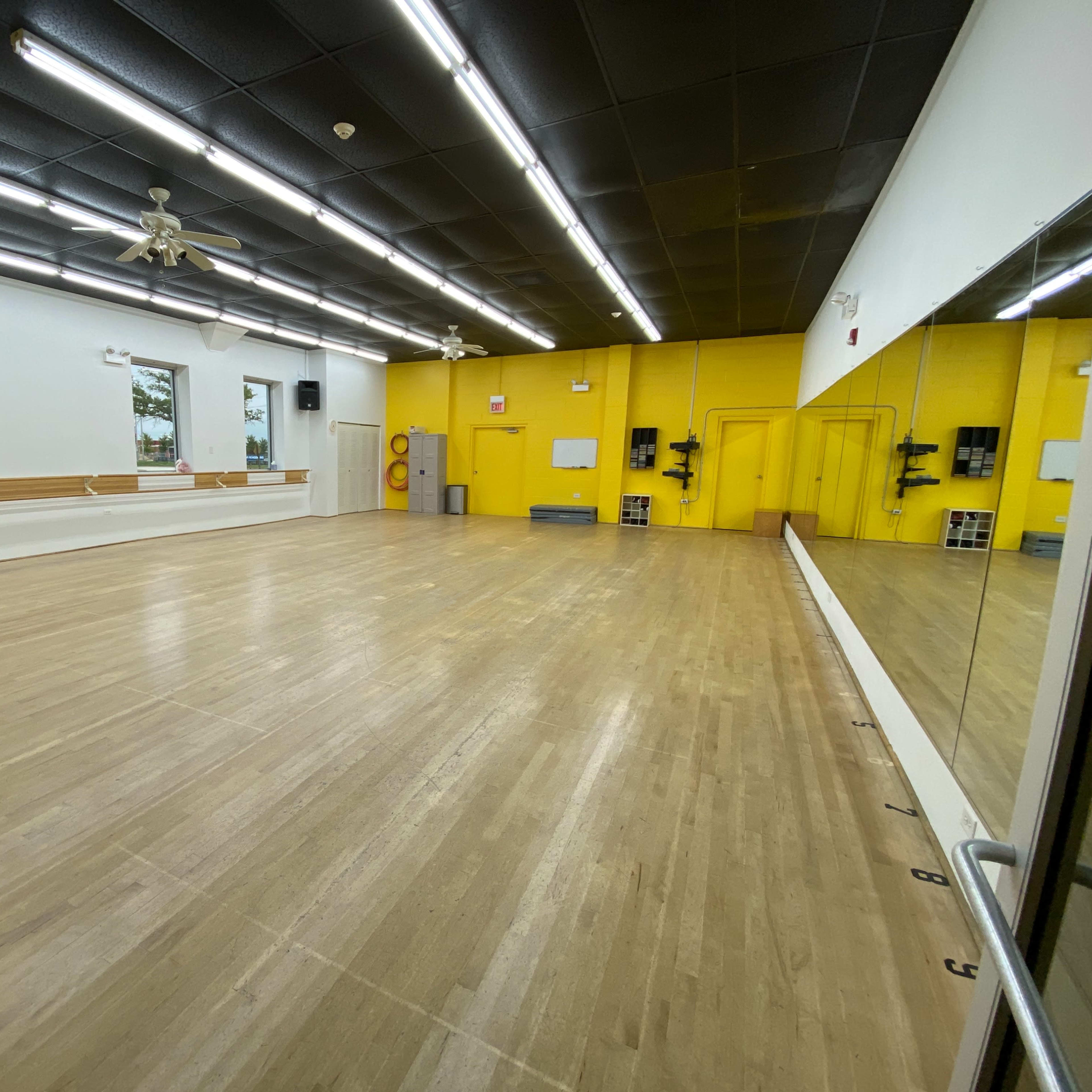 A spacious dance studio with wooden flooring, bright yellow walls, and large mirrored panels along one side.