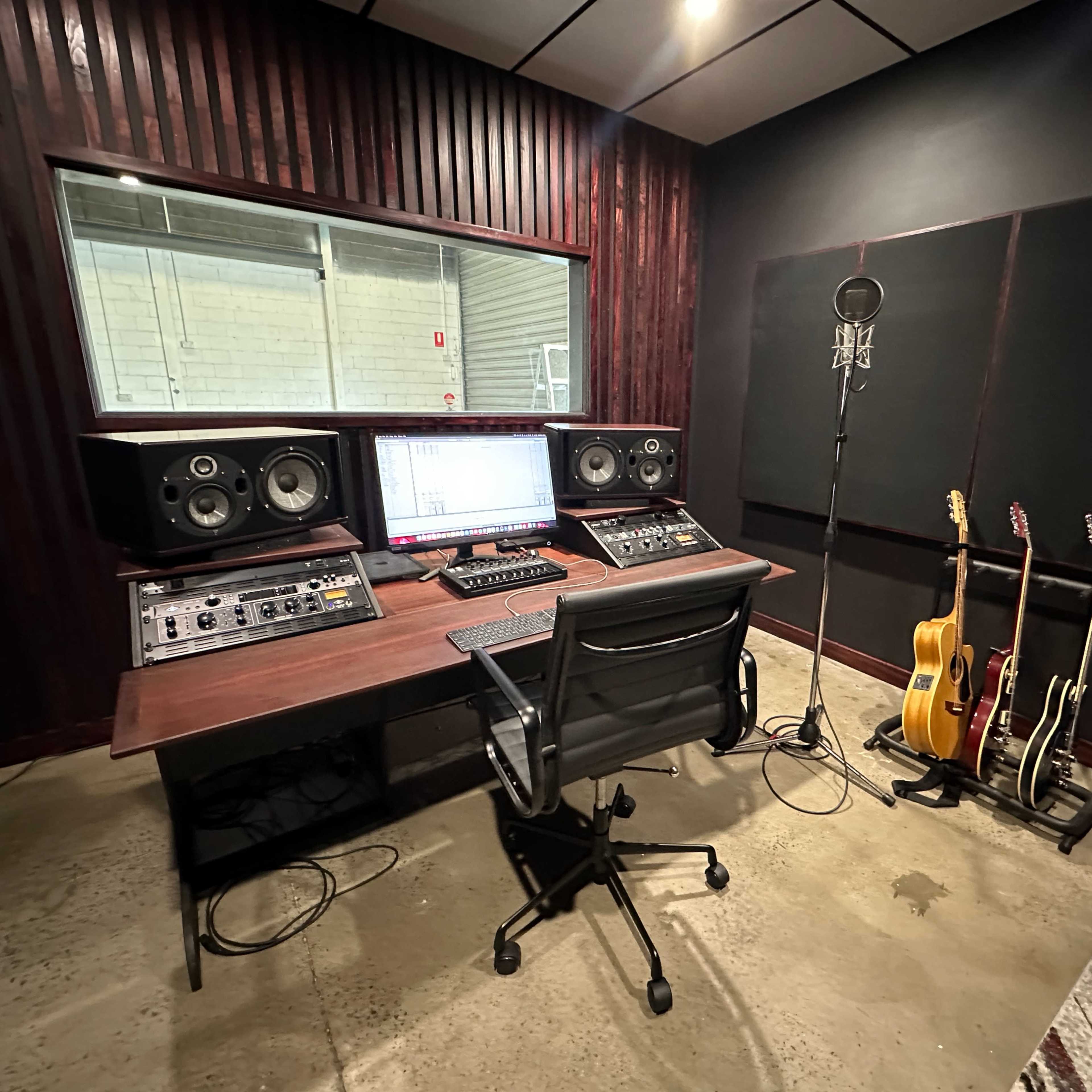 A music studio features a mixing desk with audio equipment, a computer screen, a microphone on a stand, and guitars stored in a rack.