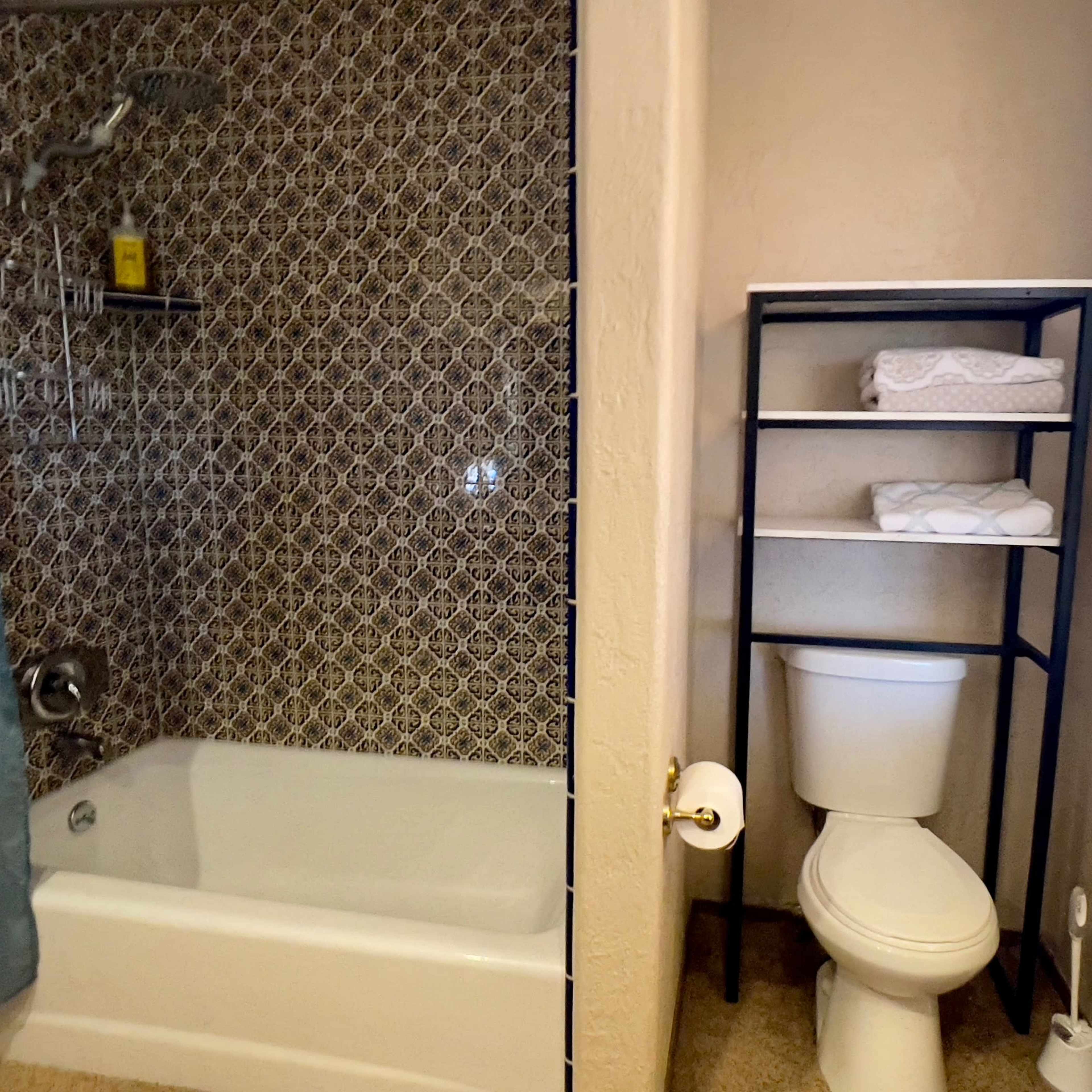 A bathroom featuring a bathtub with a shower, a toilet, and a shelving unit with neatly arranged towels.