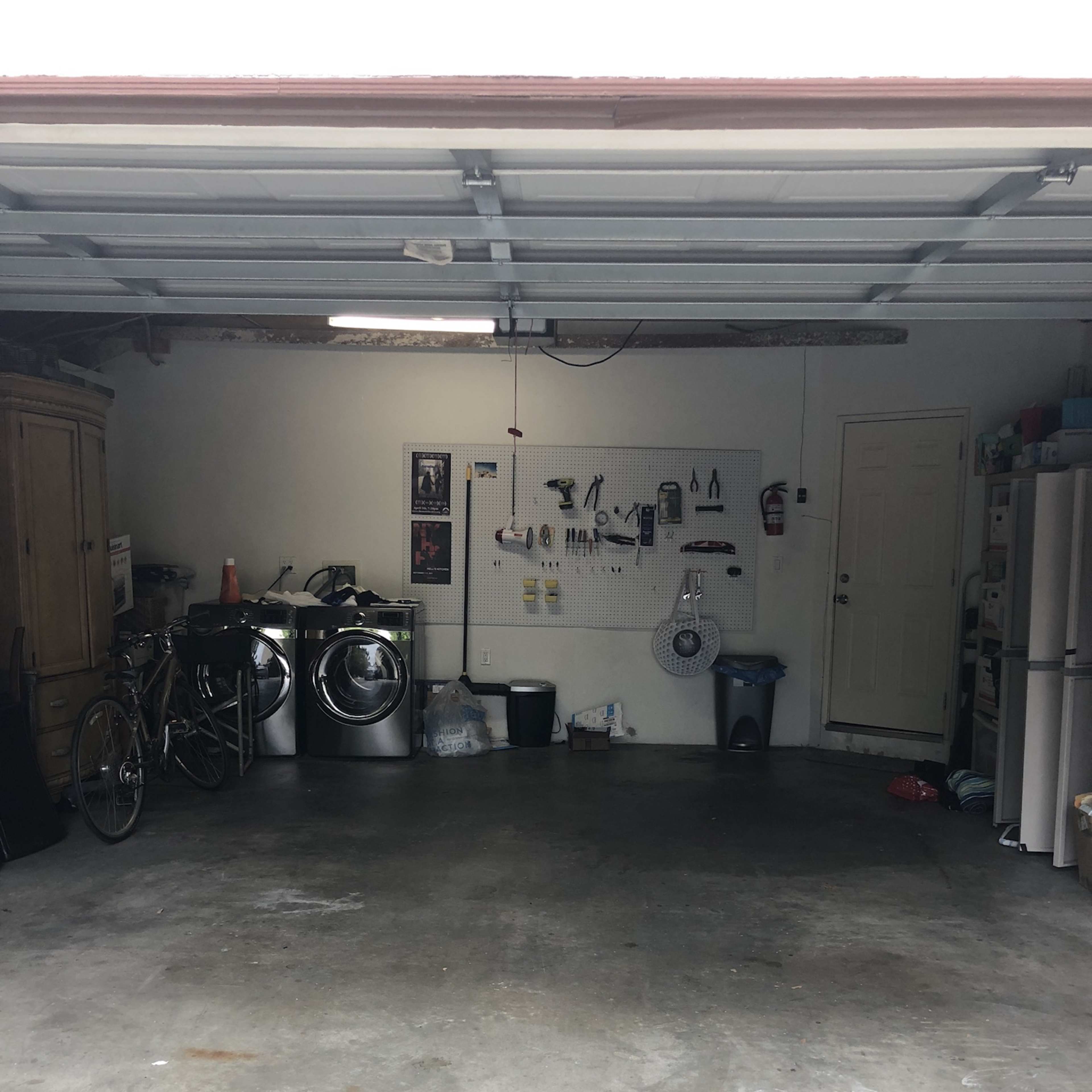 The garage contains two washing machines, a bicycle, a trash can, and various tools hanging on a wall, with a refrigerator in the back corner.