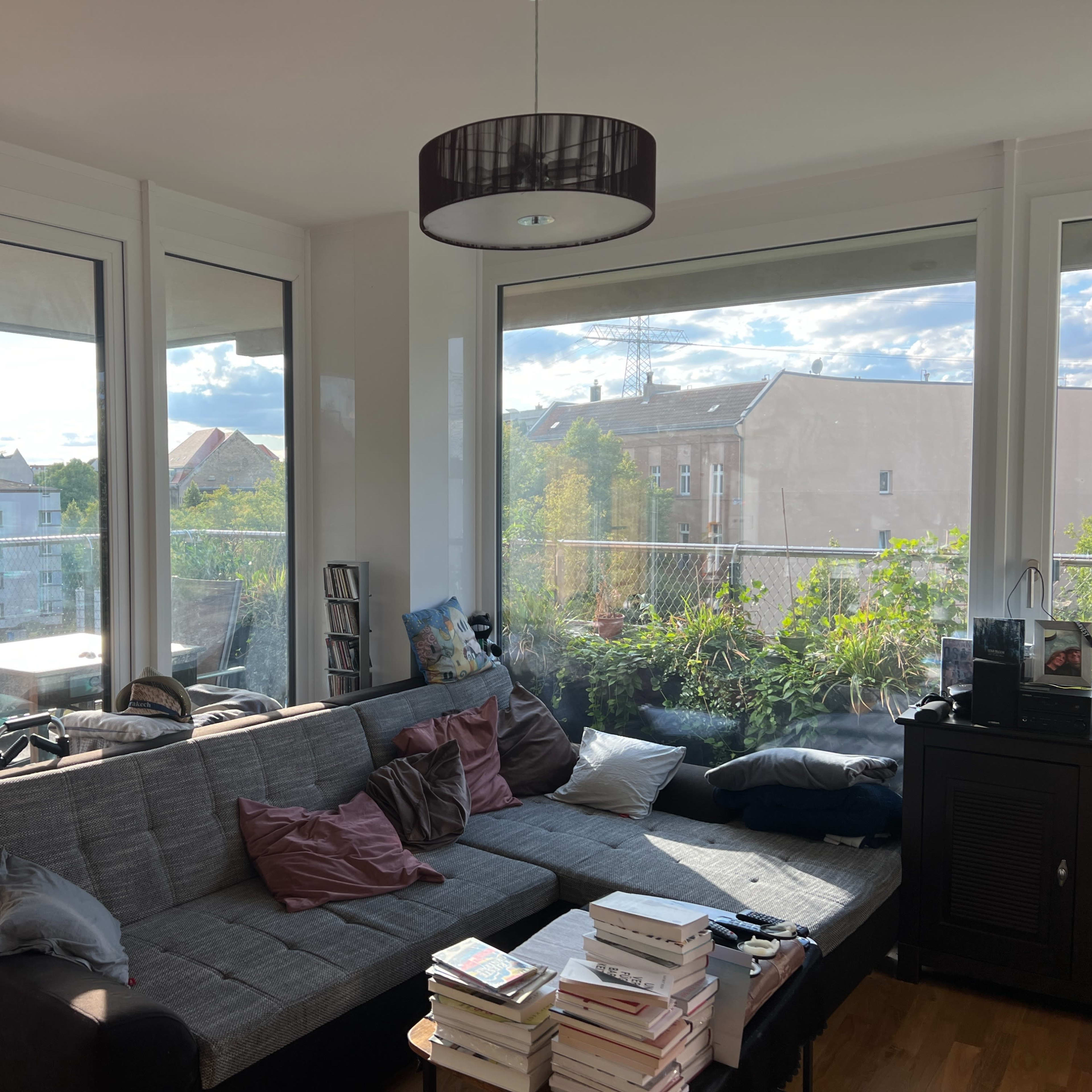 A cozy living room with large windows that overlook a balcony and greenery outside, featuring a gray sofa adorned with pillows and a coffee table stacked with books.