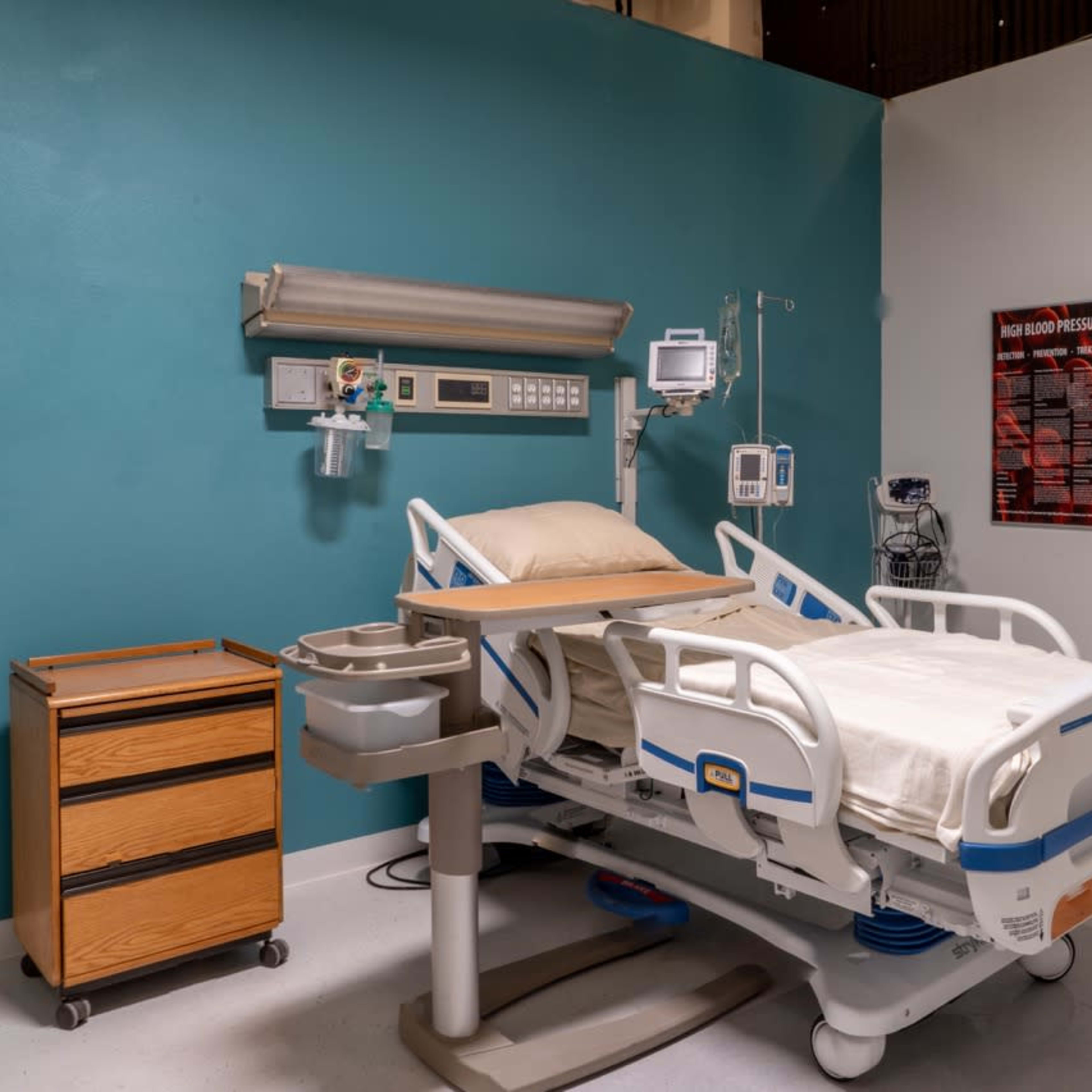 A hospital room featuring a medical bed, a mobile bedside table, and various medical equipment mounted on the wall.