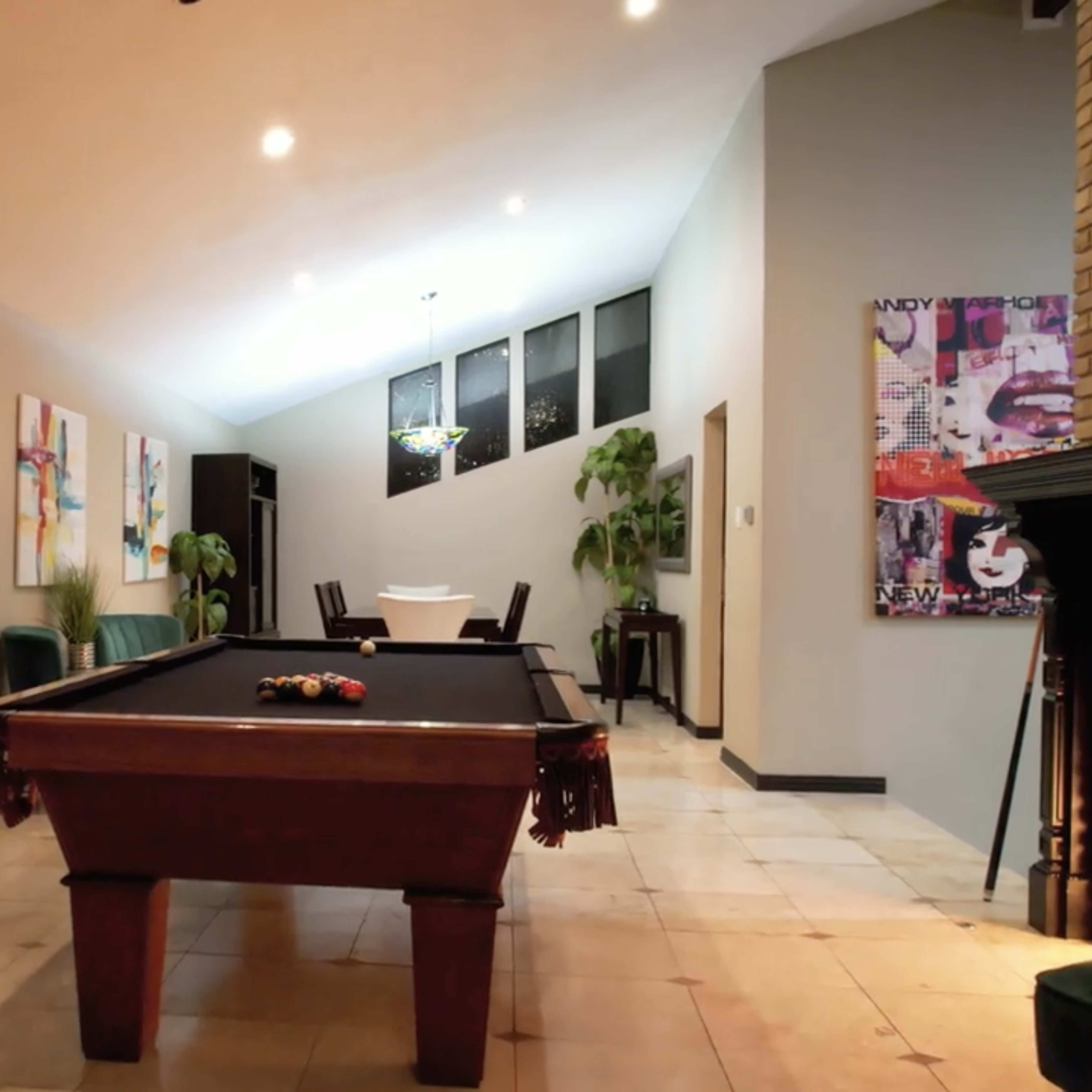 A pool table is positioned in a spacious room that features large windows, artwork on the walls, and a dining area in the background.
