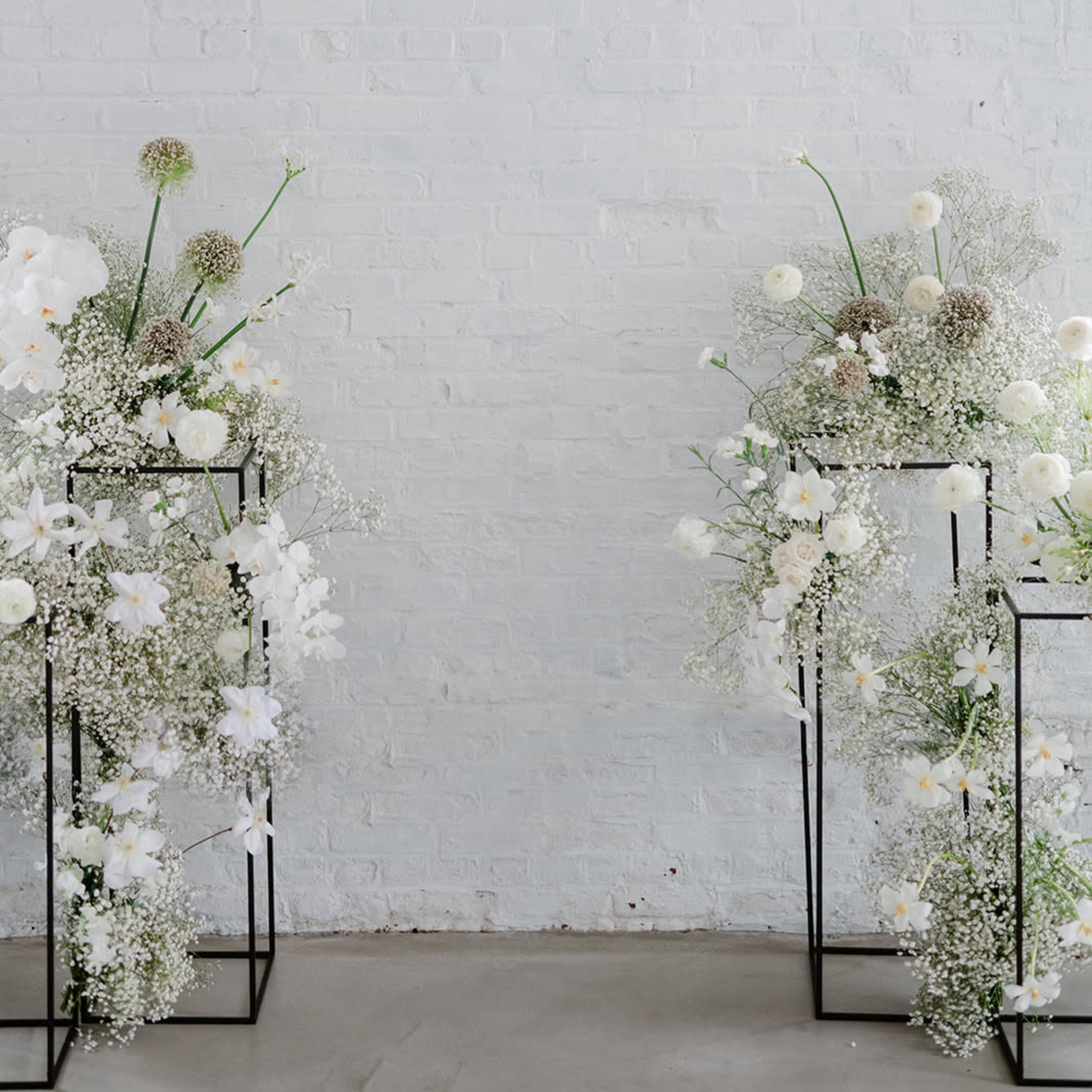 Two black metal flower stands filled with white flowers and greenery are positioned against a white brick wall.