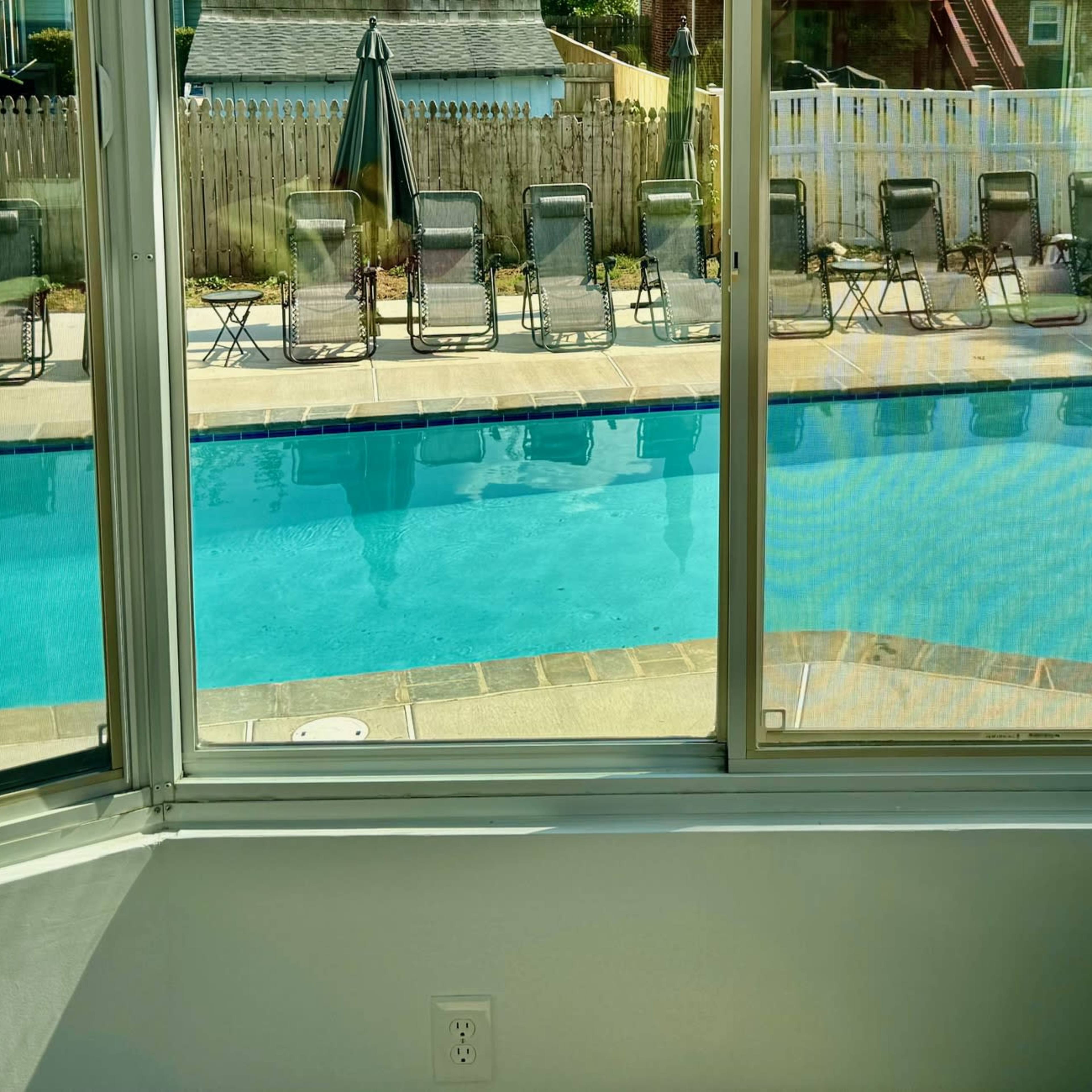 A view through a large window of a swimming pool surrounded by loungers and a patio area.