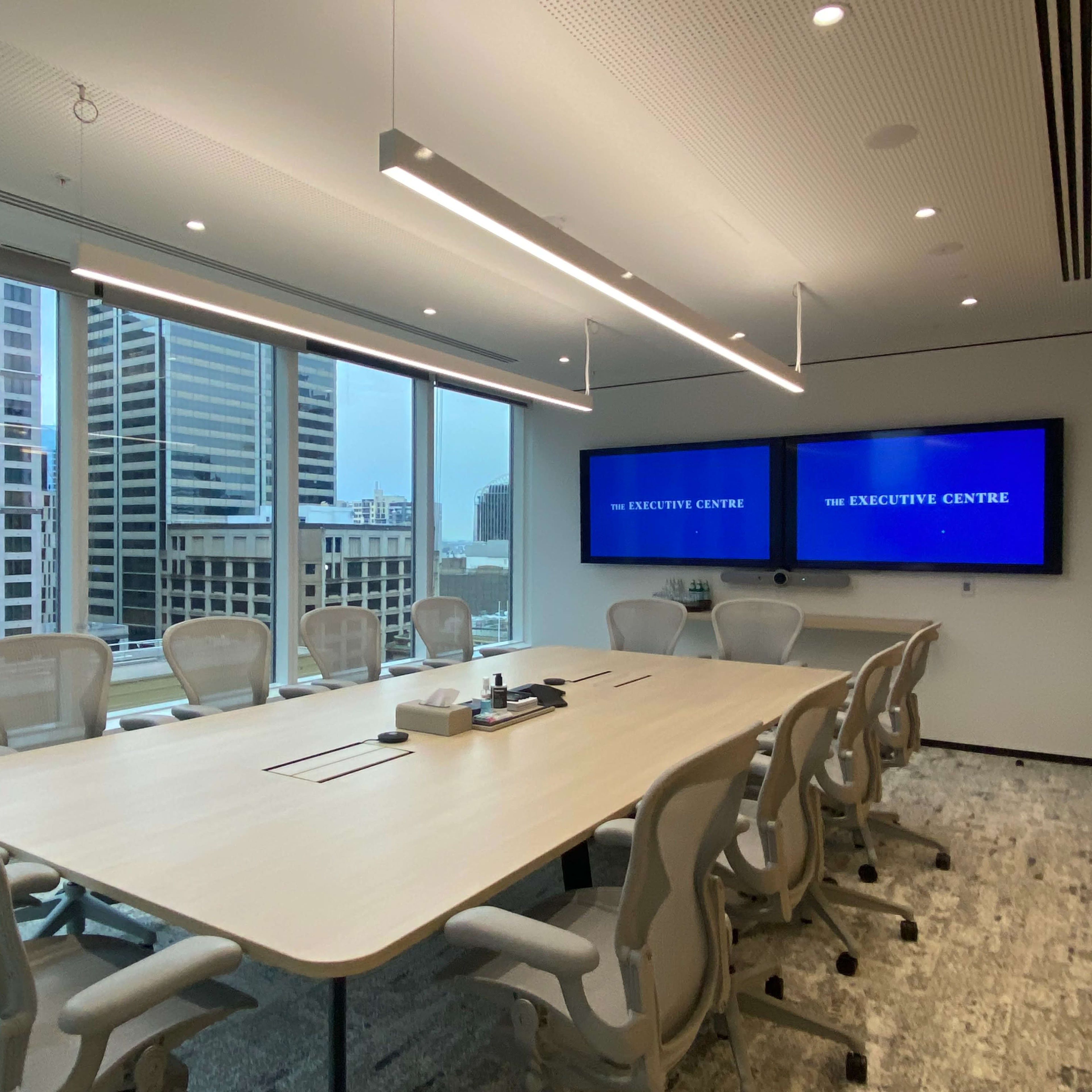 A modern conference room with a long table, ergonomic chairs, and dual screens displaying "The Executive Centre."