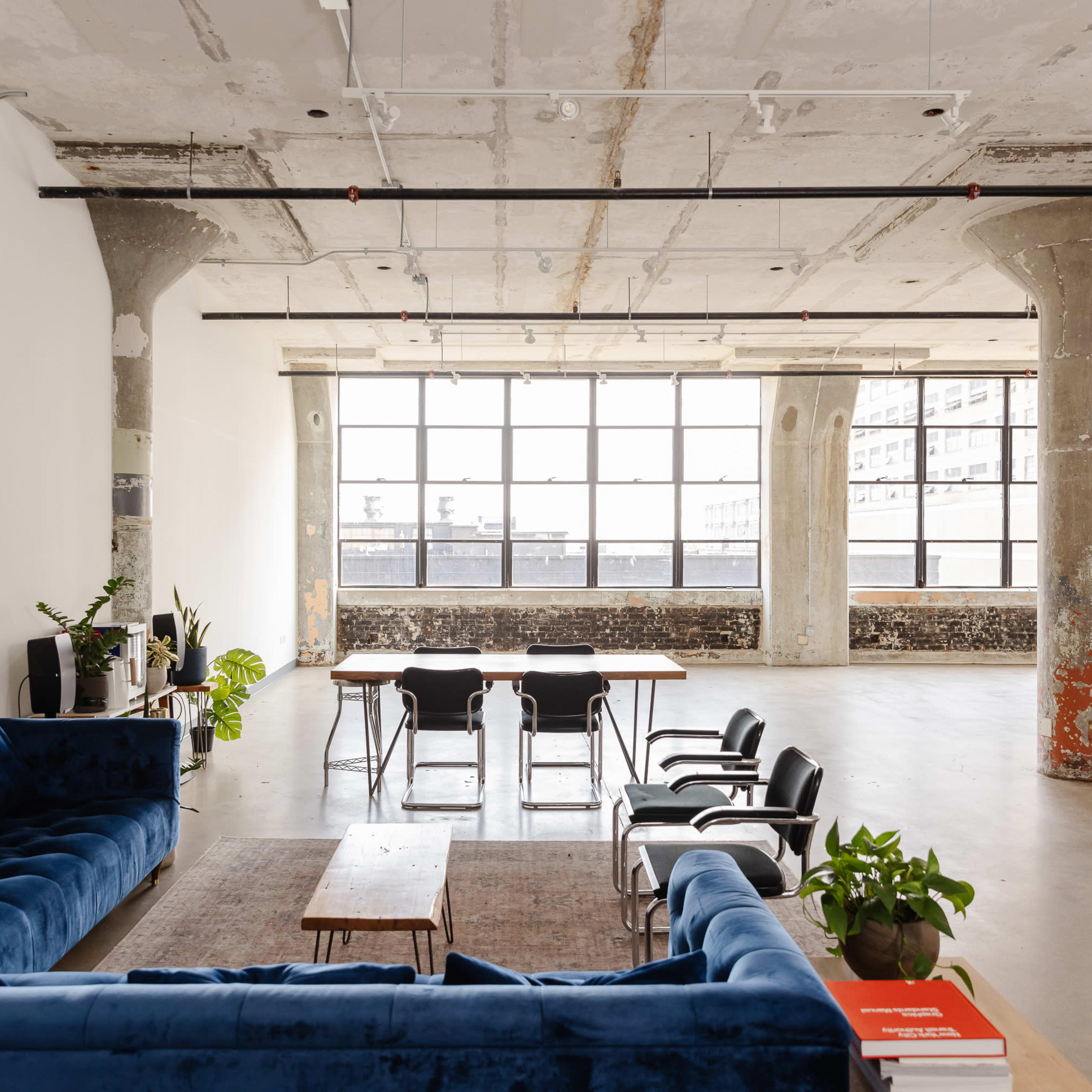 A spacious, industrial-style room features a blue sofa, a wooden coffee table, a dining table with chairs, and large windows that illuminate the space.
