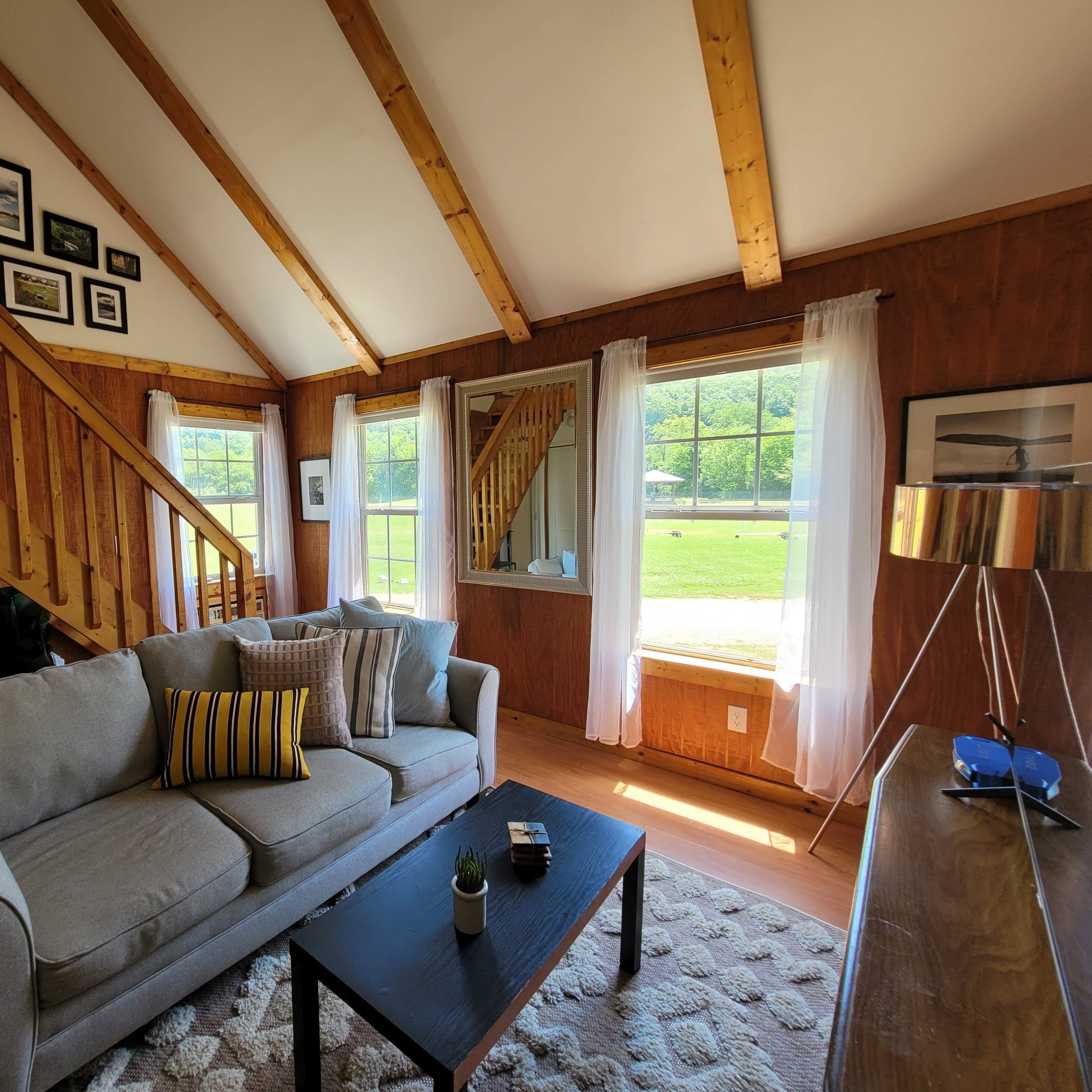 The interior of a cozy living room features a gray sofa with decorative pillows, a coffee table, and large windows that offer views of a green outdoor landscape.