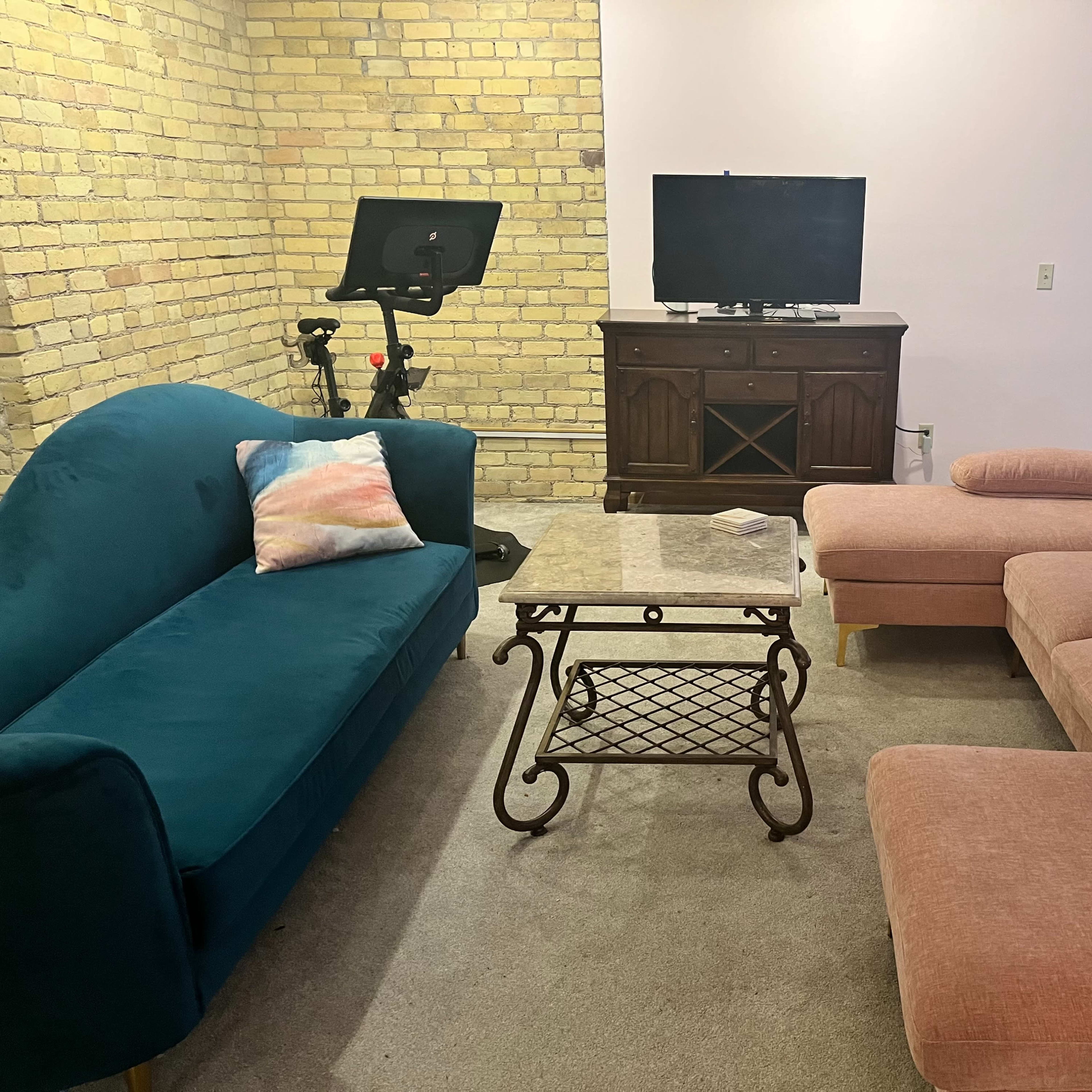 The living room features a blue sofa, a pink sectional couch, a glass-top coffee table, a television on a wooden cabinet, and an exercise bike.
