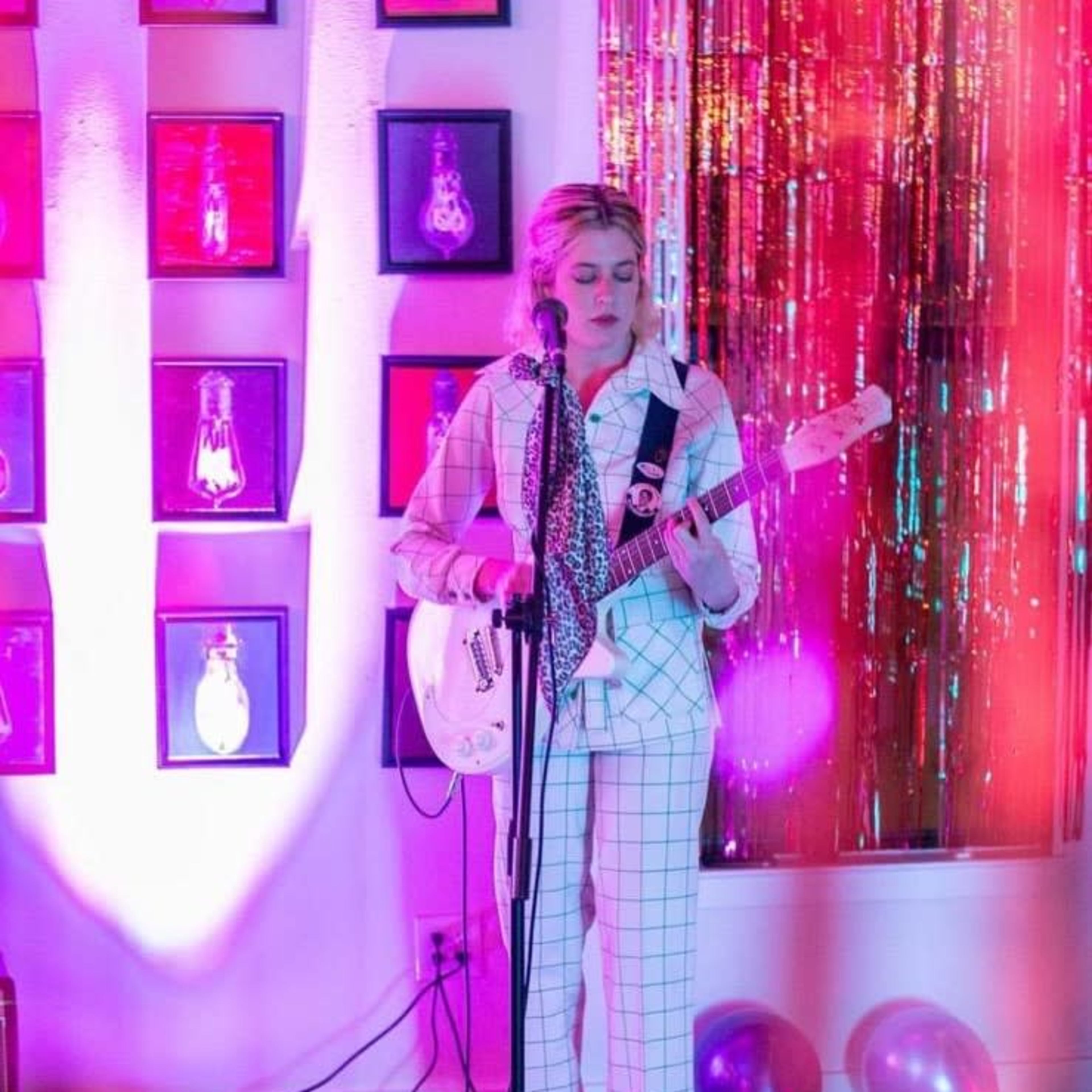 A musician stands on stage with a guitar, surrounded by colorful lights and decorations, as balloons lie on the floor.