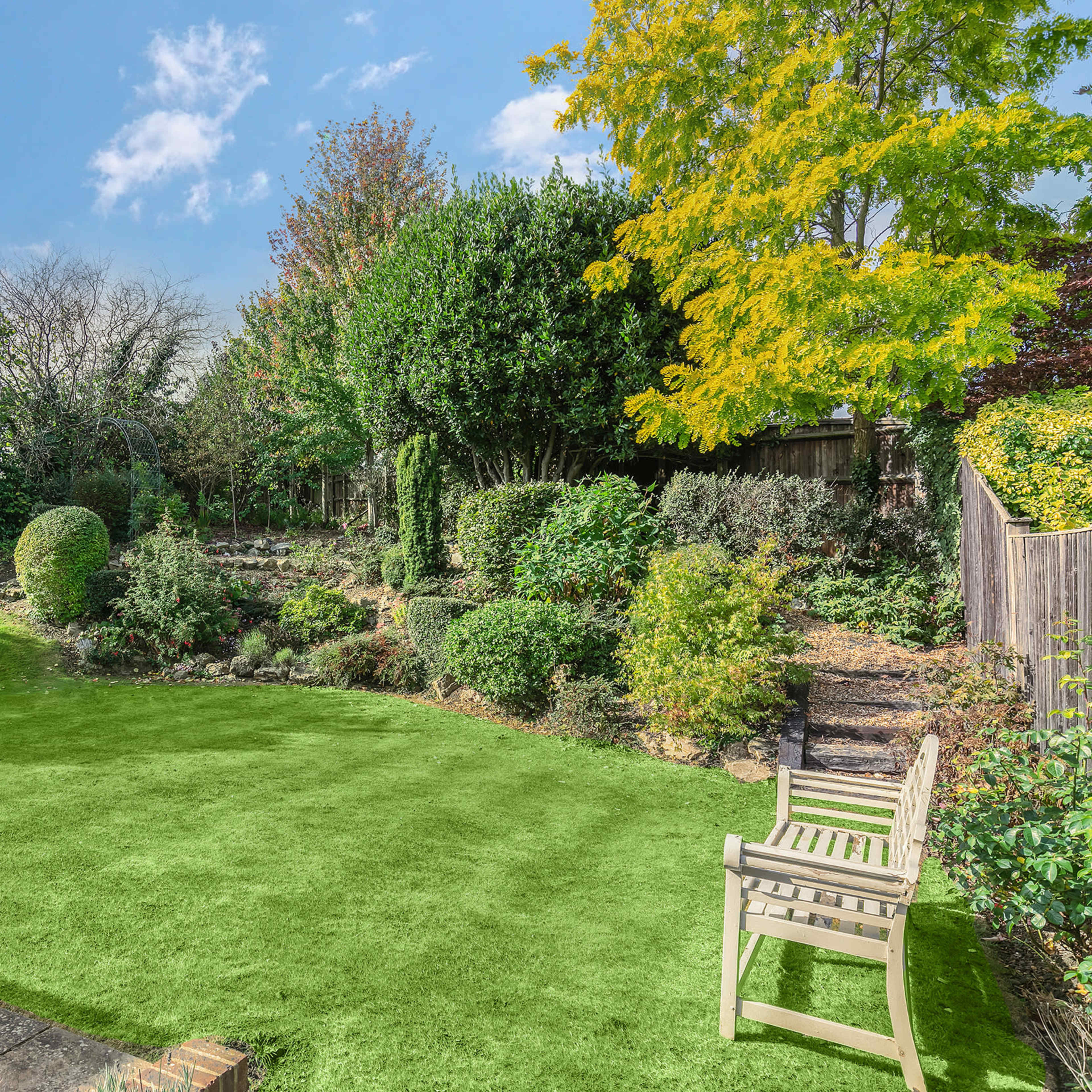 A well-maintained garden with lush greenery, a pond, and a wooden bench in a sunlit setting.