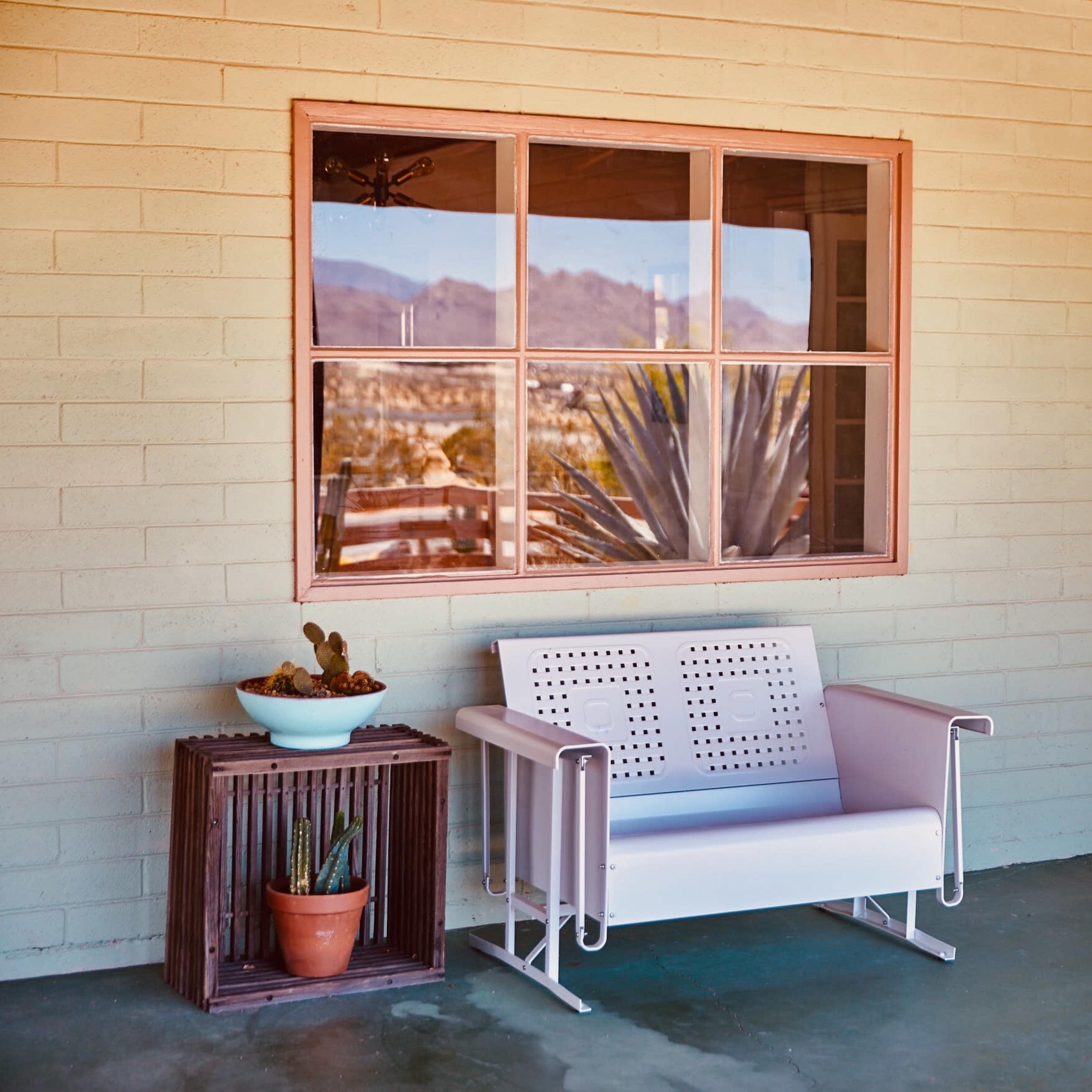 A porch area featuring a white bench, a small plant pot, and a door with a window reflecting mountains in the background.