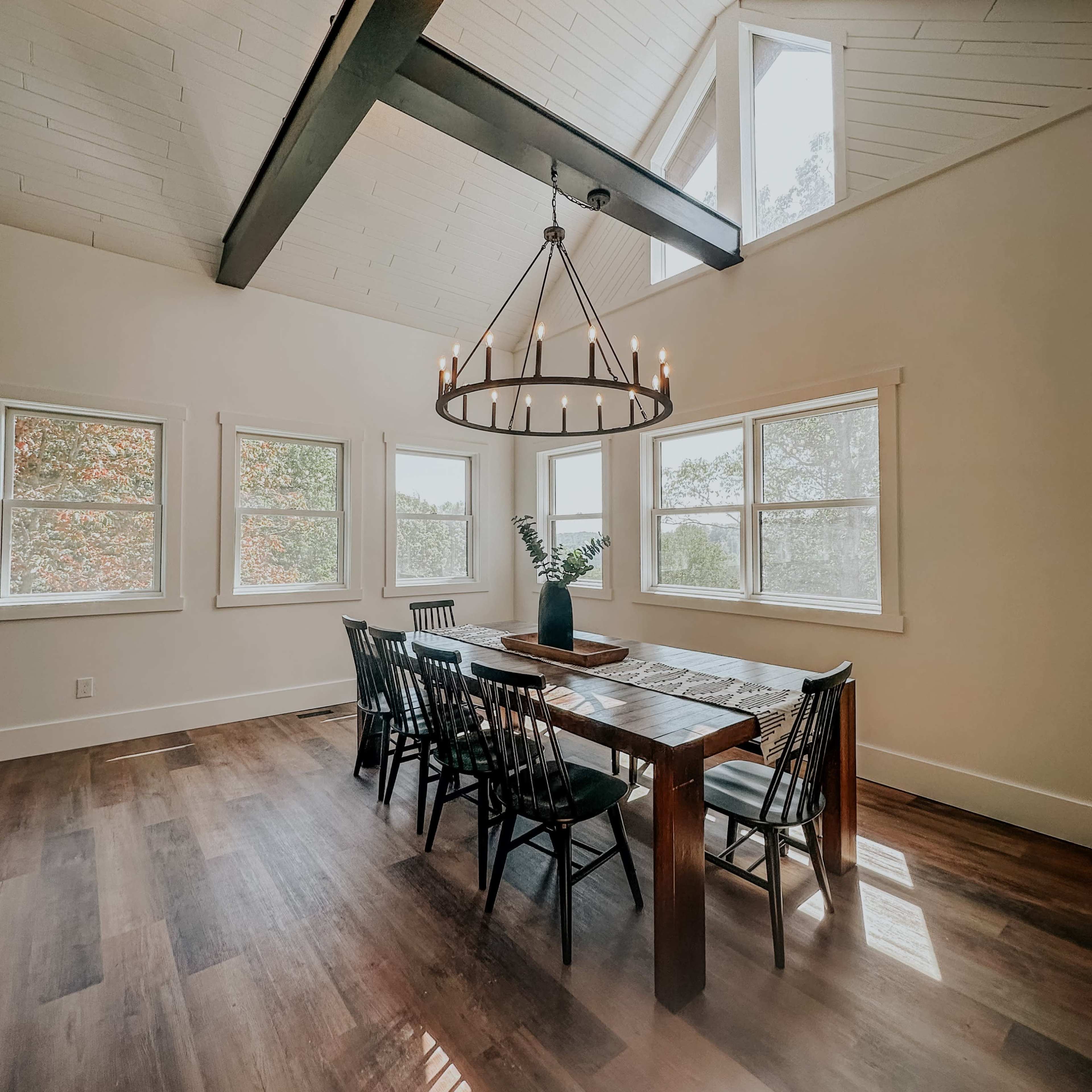 A spacious dining area features a large wooden table surrounded by black chairs beneath a chandelier, with several windows allowing natural light to fill the room.