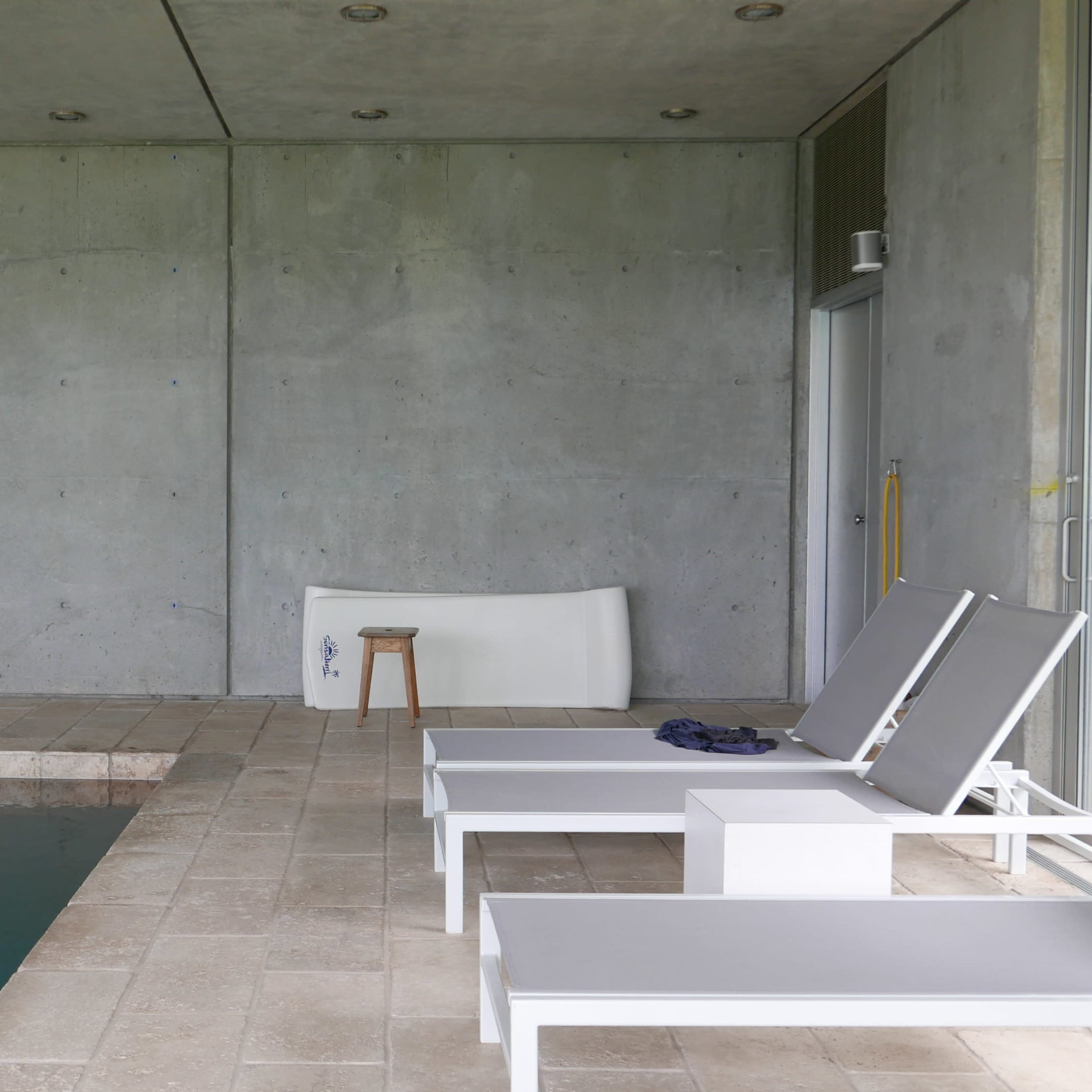 The scene features a modern indoor pool area with gray concrete walls, a tiled floor, and two white lounge chairs beside the water.